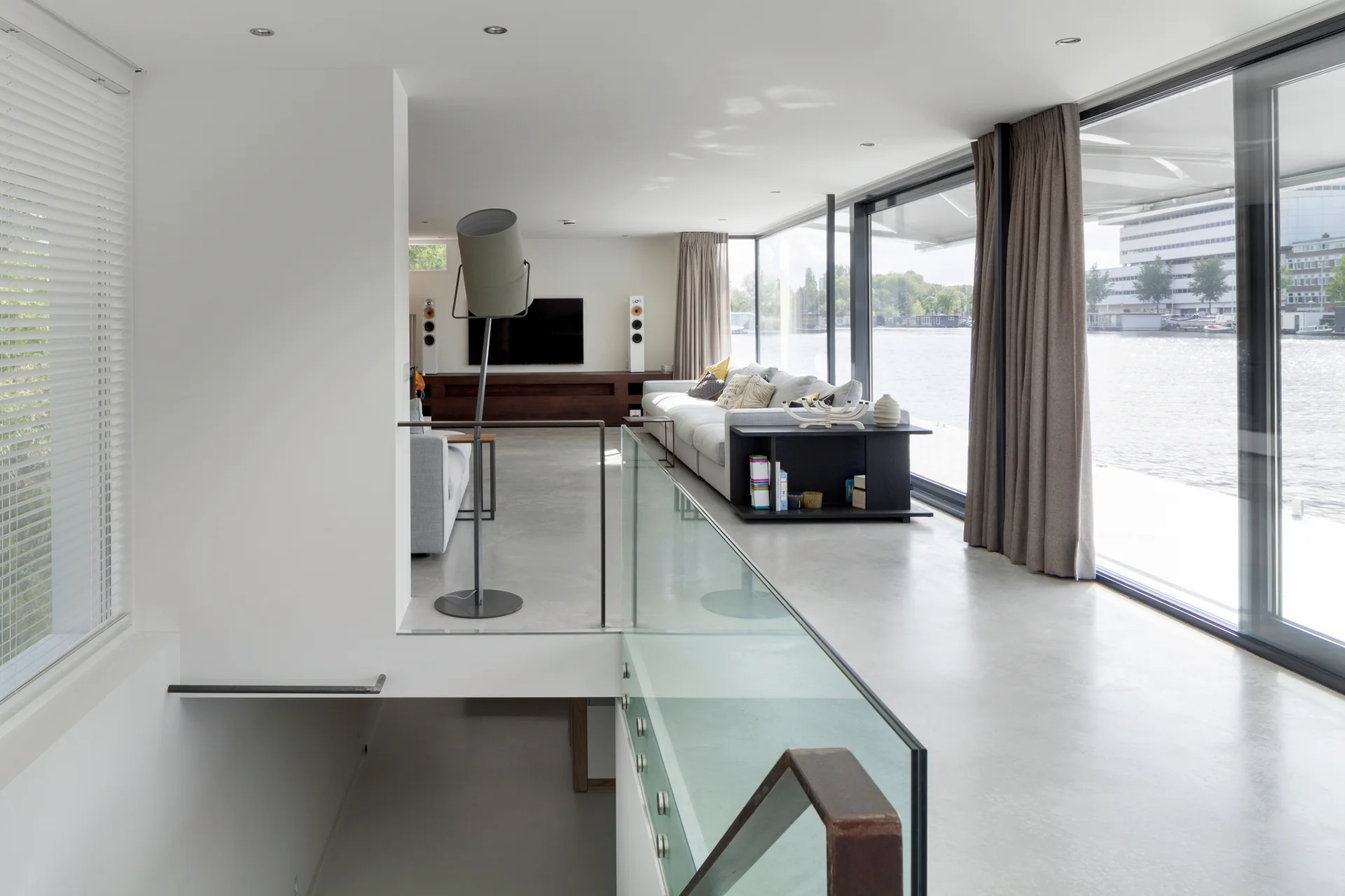 A modern split-level living room features a grey sofa, glass staircase railing, concrete-like flooring, and large windows with water views.