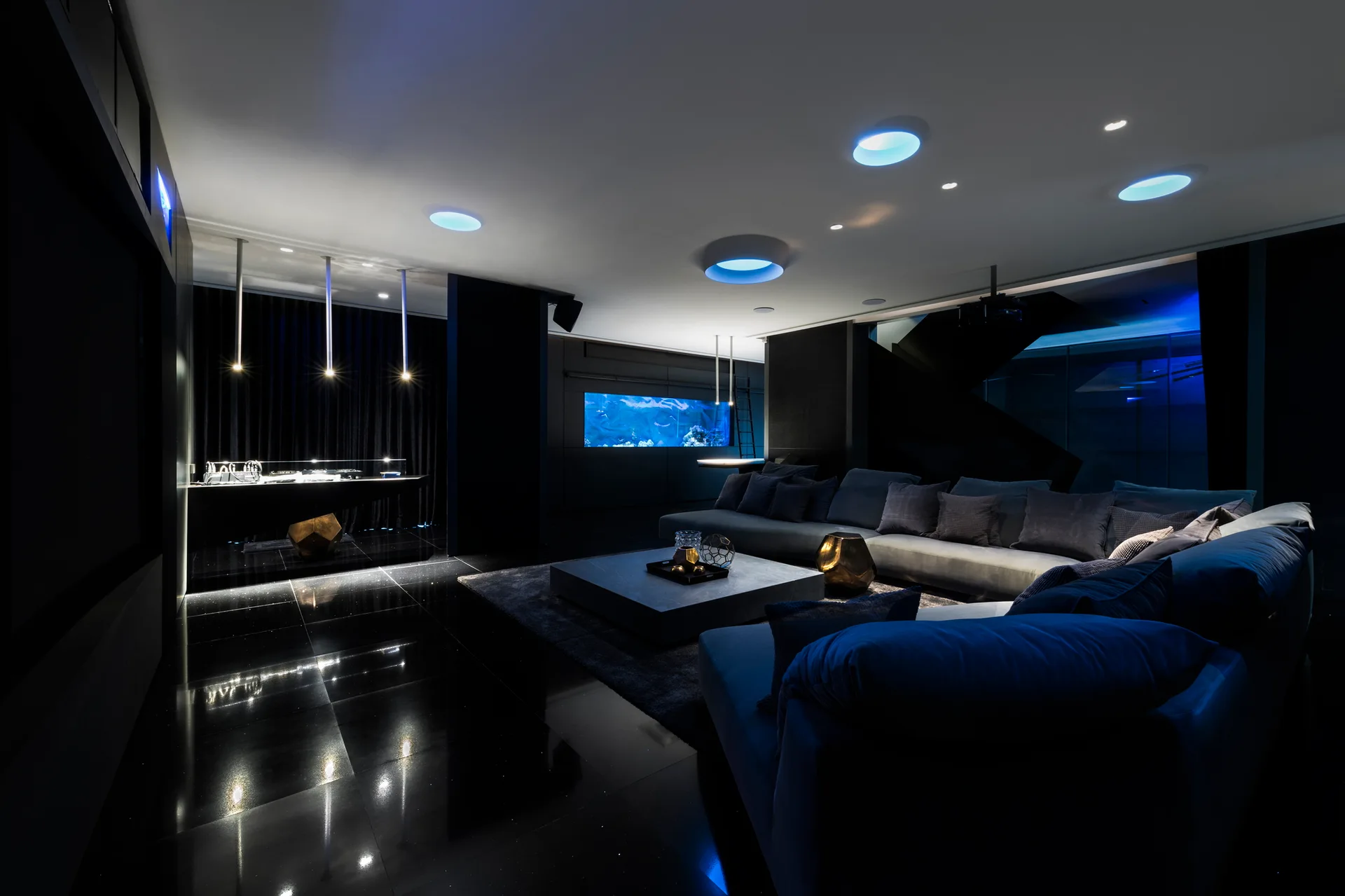 A dark media room features a grey sectional sofa, a built-in wall aquarium, and blue circular recessed ceiling lights.