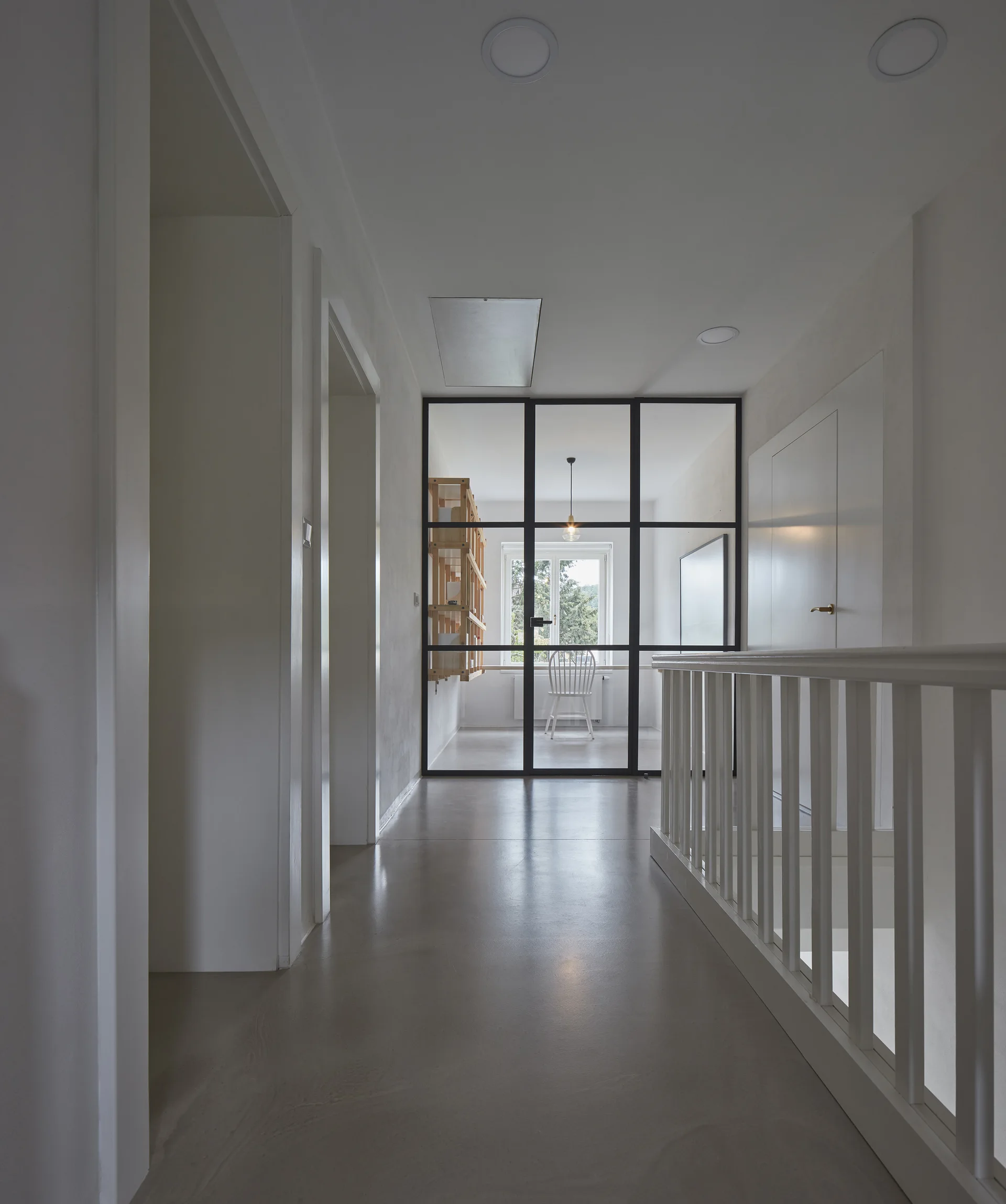 A minimalist white hallway features a grey floor, a white stair railing, and a black steel-framed glass partition wall leading to a sunlit room.