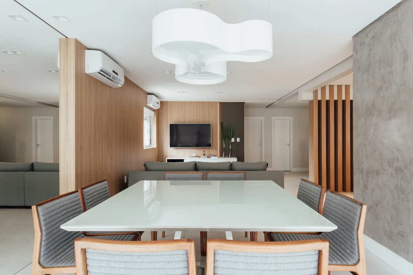 A modern dining room features a white-topped table with grey upholstered chairs beneath a large white sculptural pendant light and wood-paneled walls.
