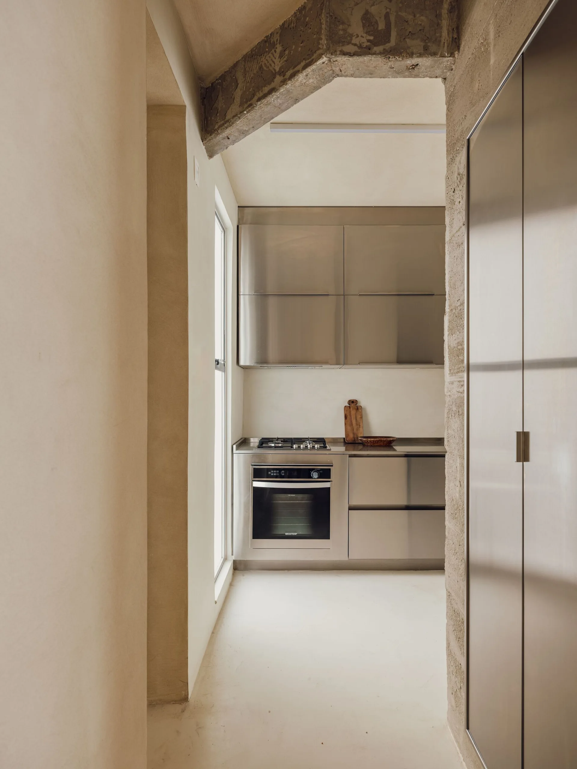A minimalist kitchen features stainless steel cabinetry and built-in appliances framed by warm plaster walls and an exposed concrete beam.