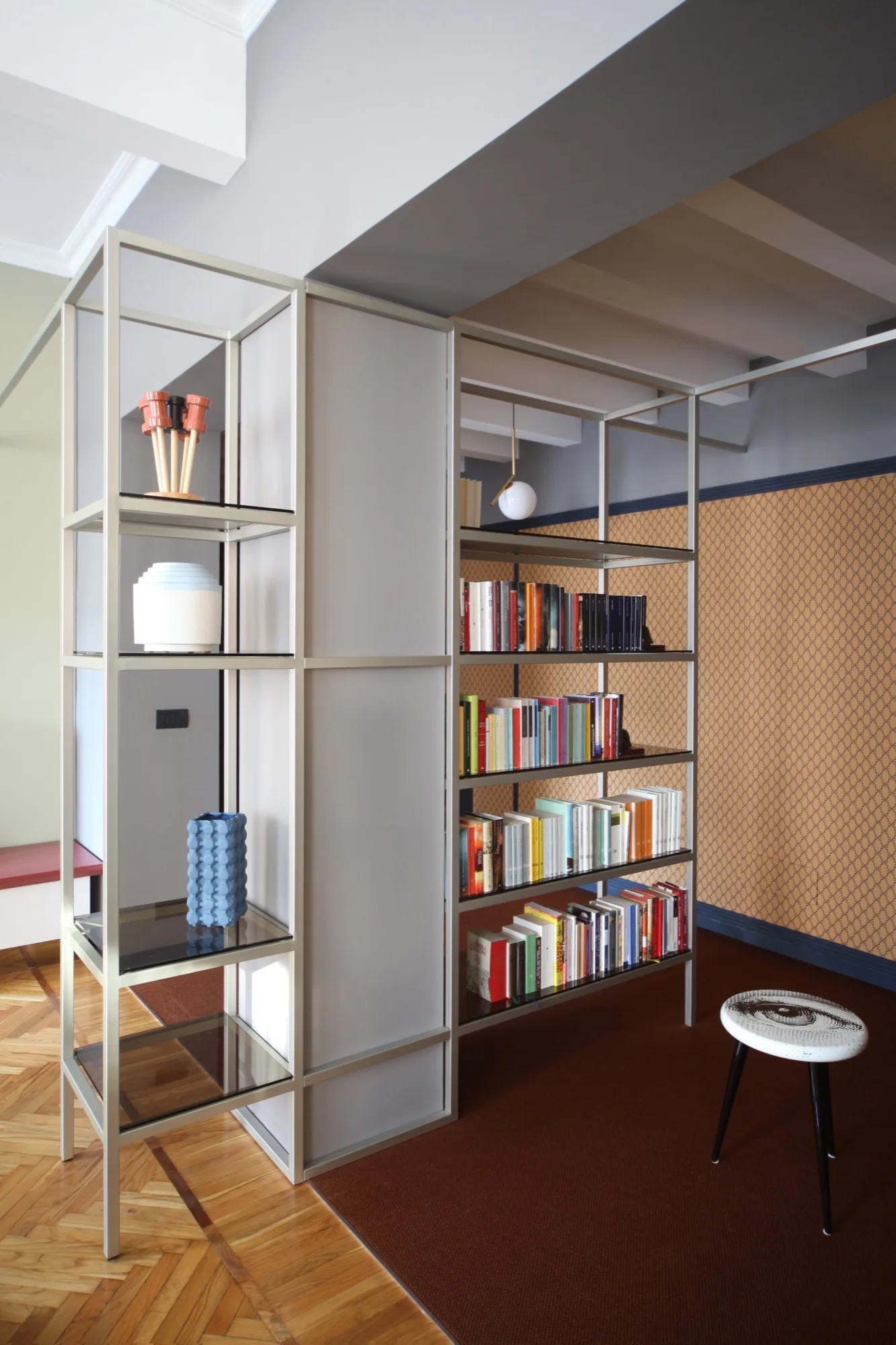 A silver metal shelving unit serves as a room divider, holding books and decor objects on herringbone parquet wood flooring.