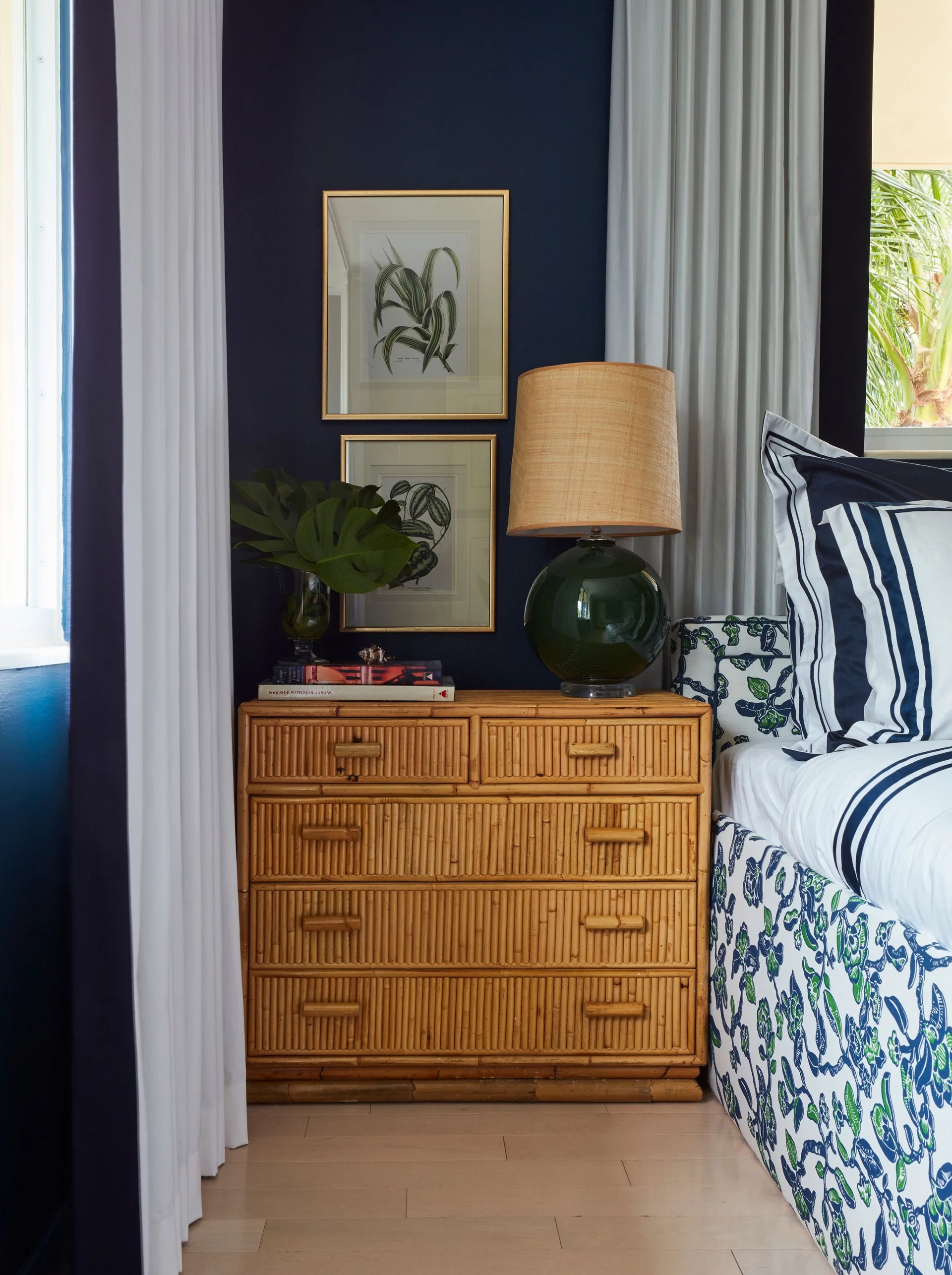 A bamboo chest of drawers sits against a dark navy blue wall next to a bed, topped with a green glass lamp and botanical artwork.