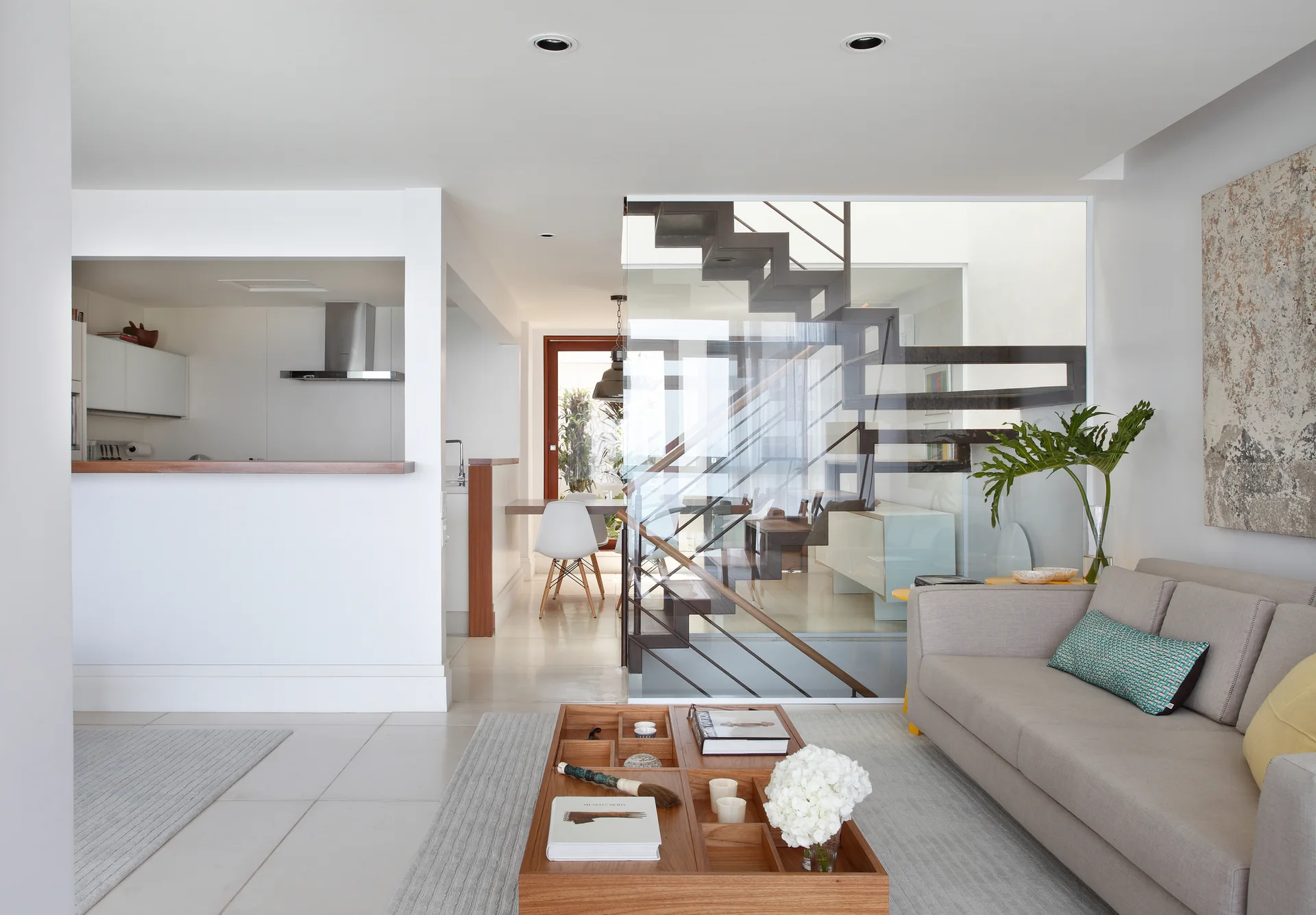 A contemporary open-plan living room with a grey sofa, a wooden compartment coffee table, and a glass-enclosed floating staircase.