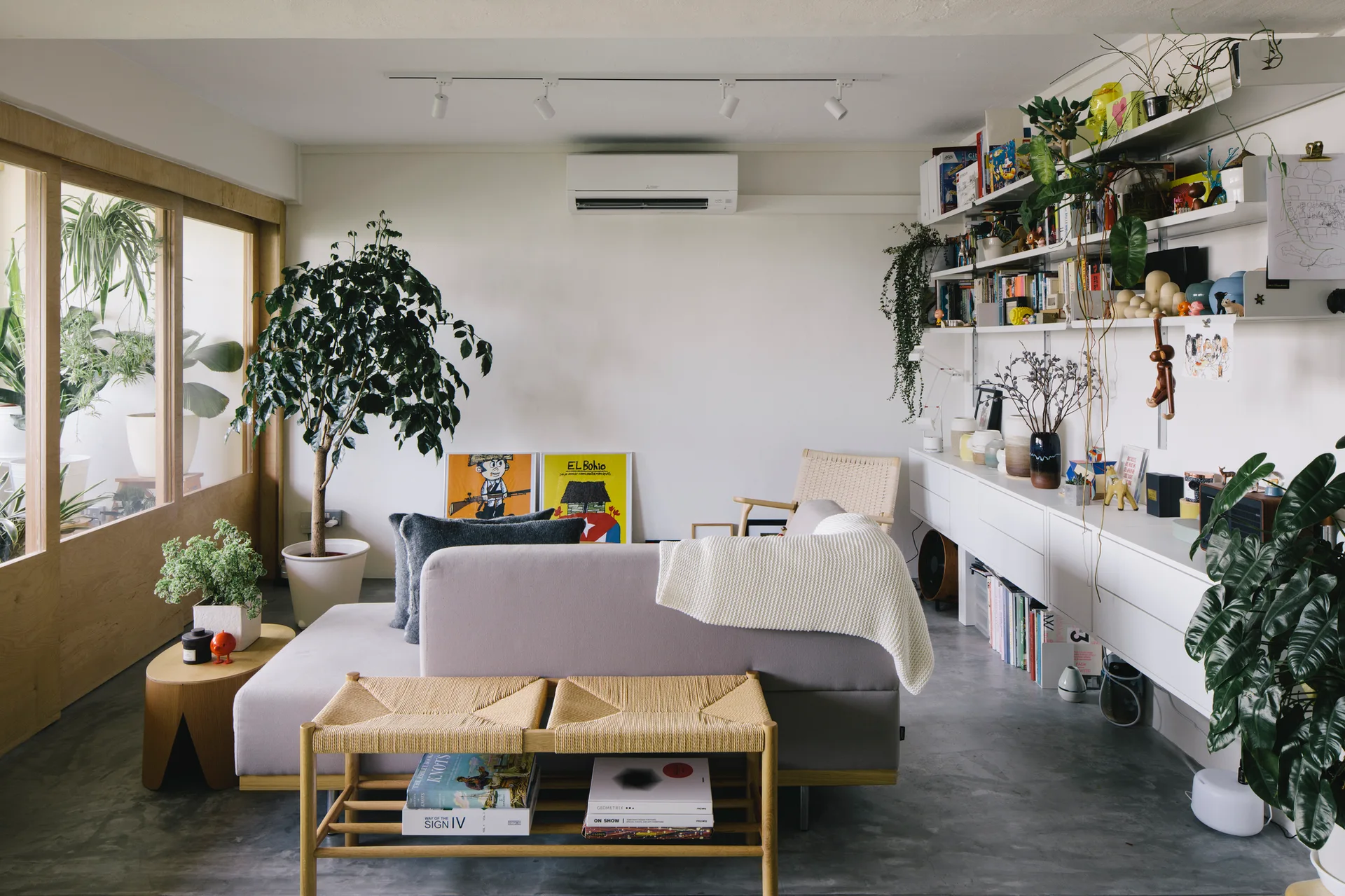 A gray sofa and a woven bench sit on a concrete floor in a light-filled living room featuring white modular wall shelving.
