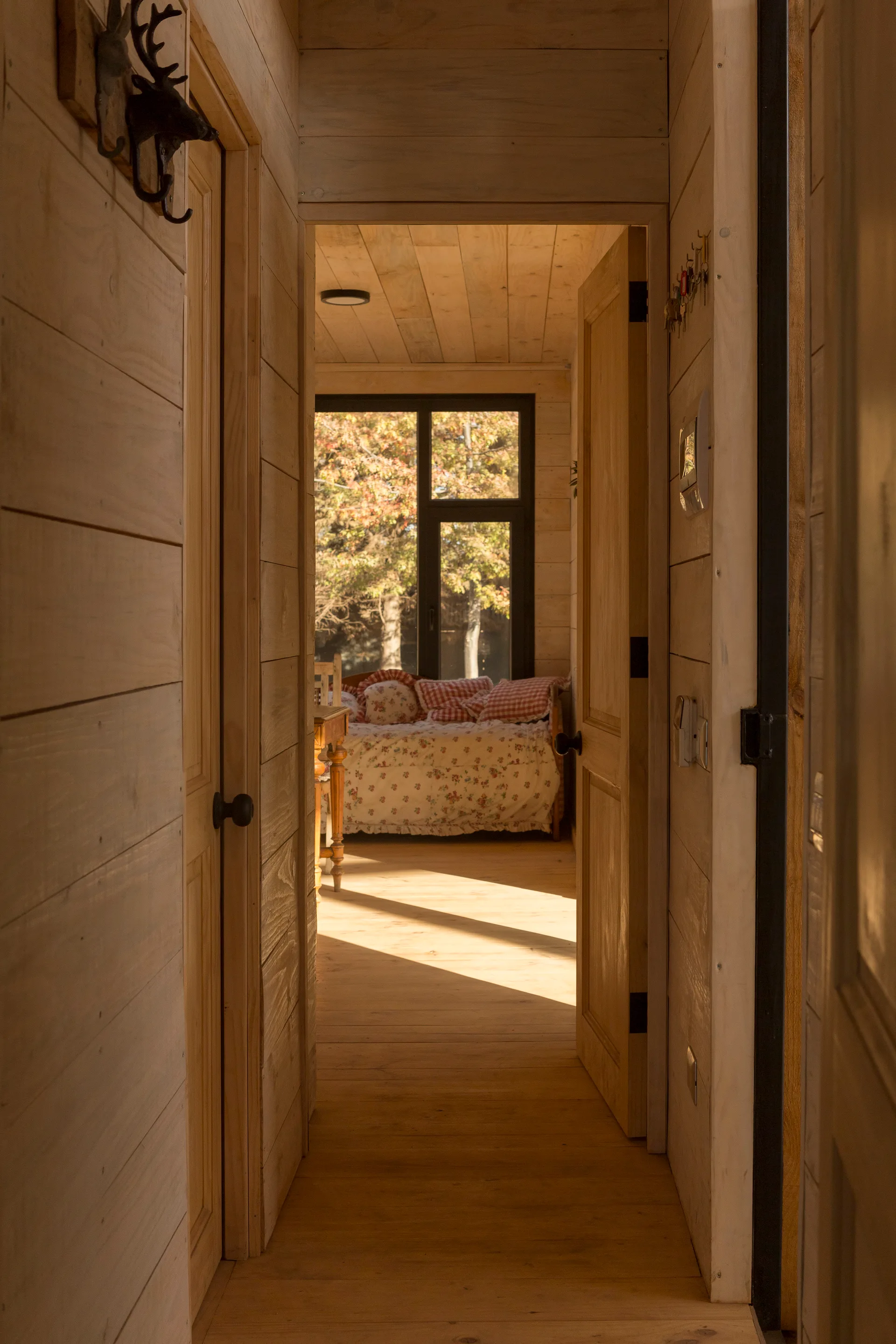 A narrow hallway with light wood plank walls and doors leads to a sunlit bedroom featuring a daybed with patterned pillows under a large window.