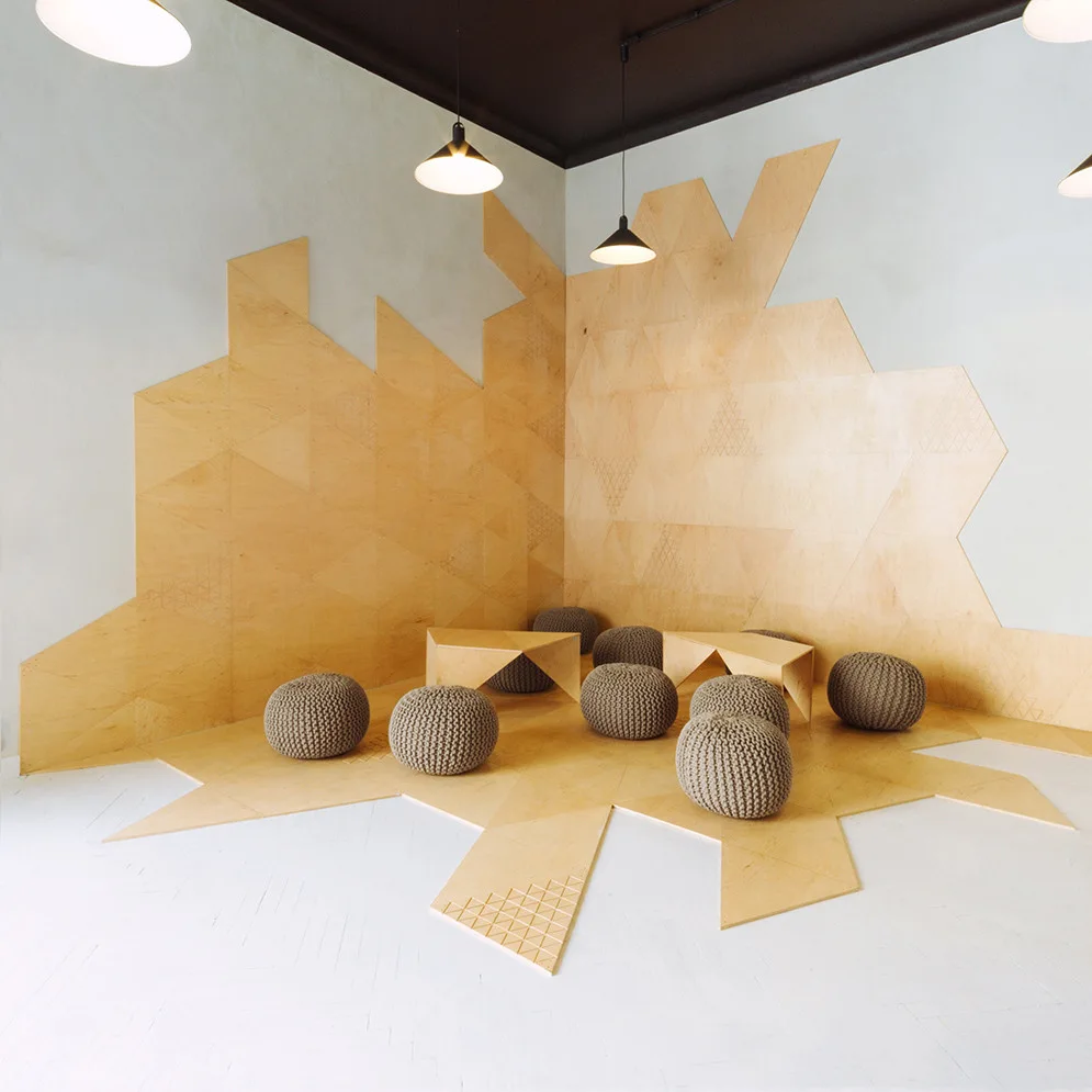 A minimalist seating area with geometric wood panels on the wall and floor, grey knitted poufs, and low triangular tables.