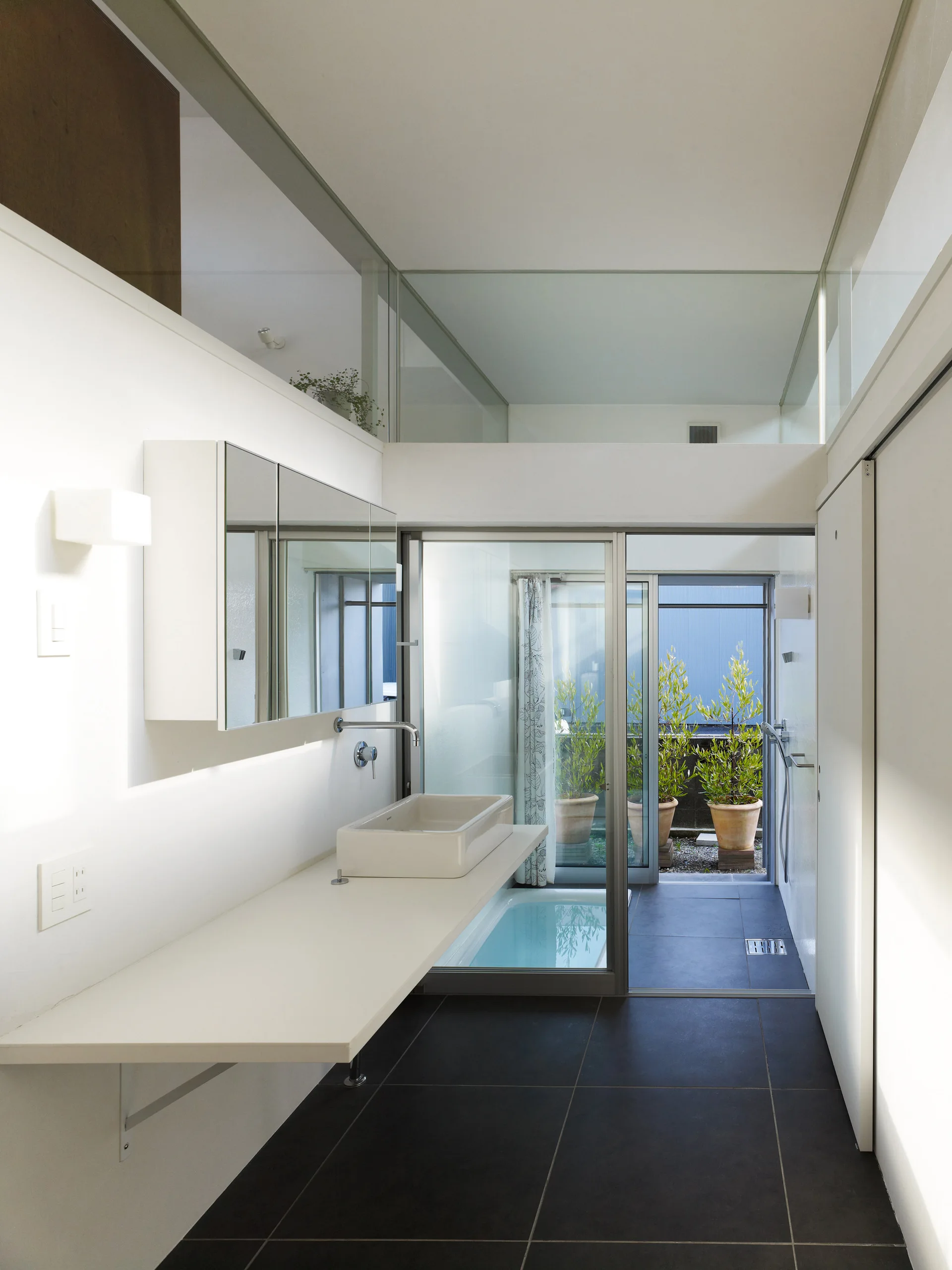 A minimalist white bathroom features a floating vanity with a vessel sink, dark floor tiles, and a sliding glass door opening to a plant-filled courtyard.