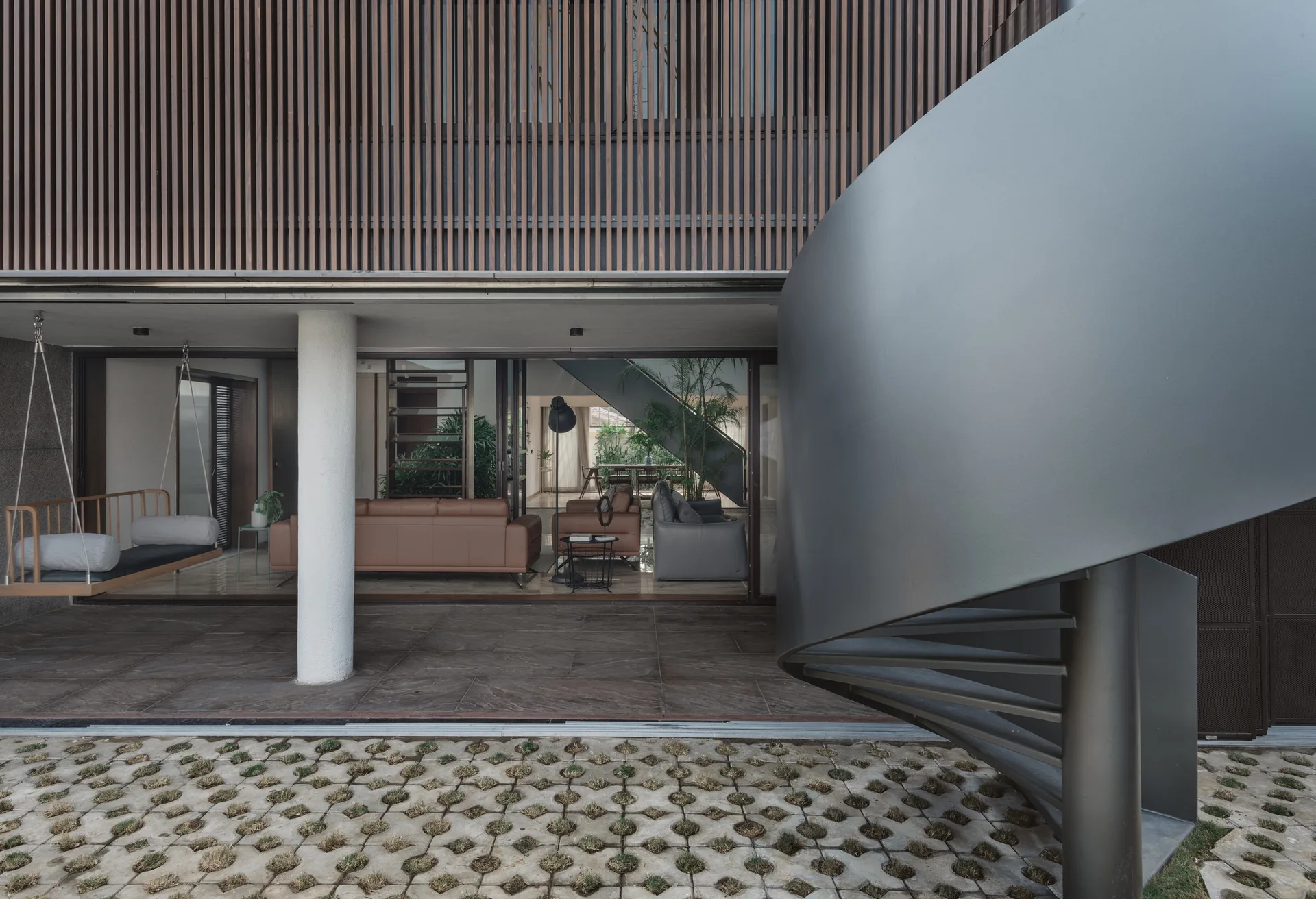 A modern covered patio features a dark metal spiral staircase, a wooden hanging swing, and sliding glass doors that open to an indoor living room.