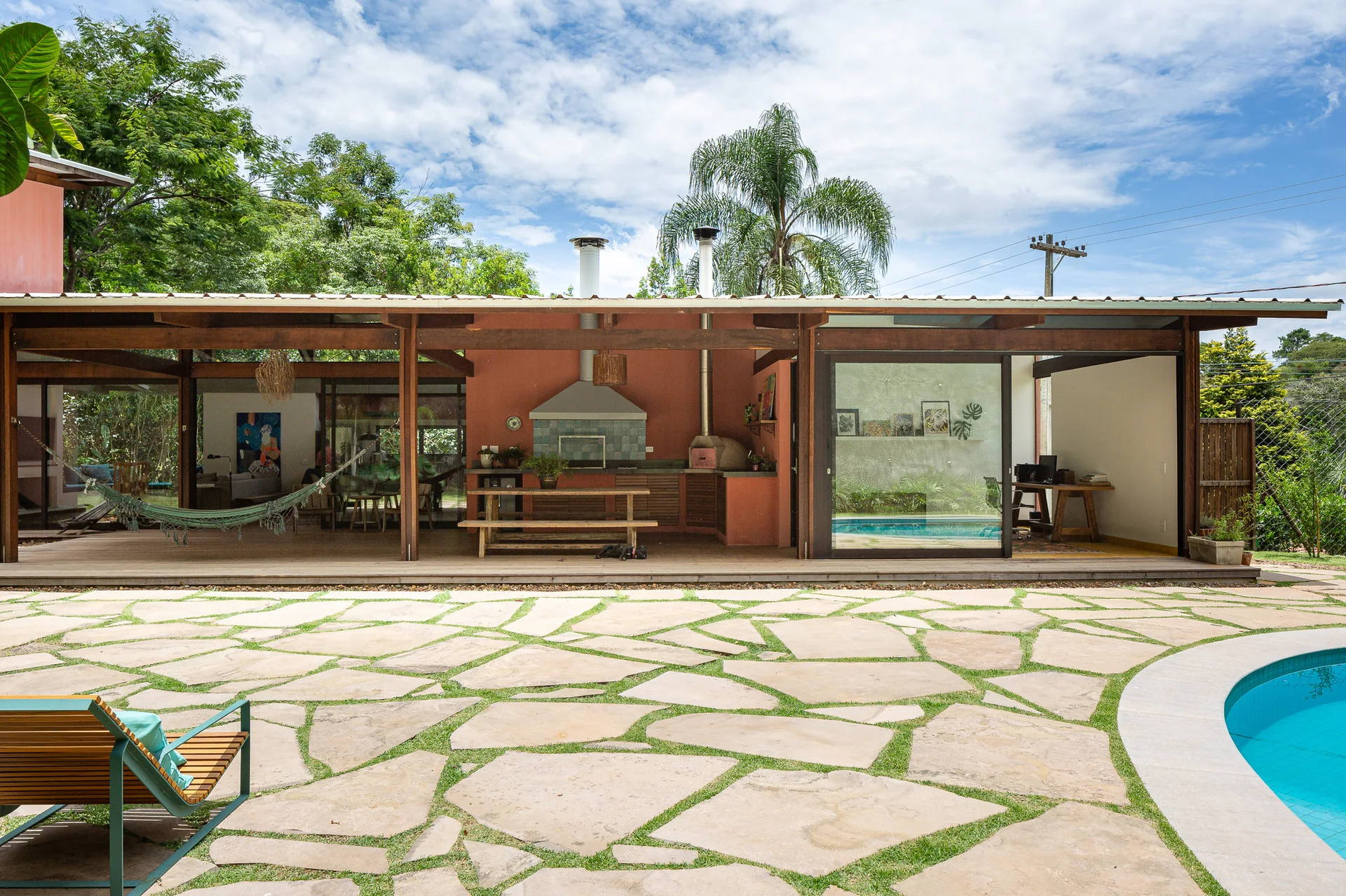 A covered outdoor patio features a wooden dining table and terracotta kitchen wall next to a flagstone pool deck with a swimming pool.