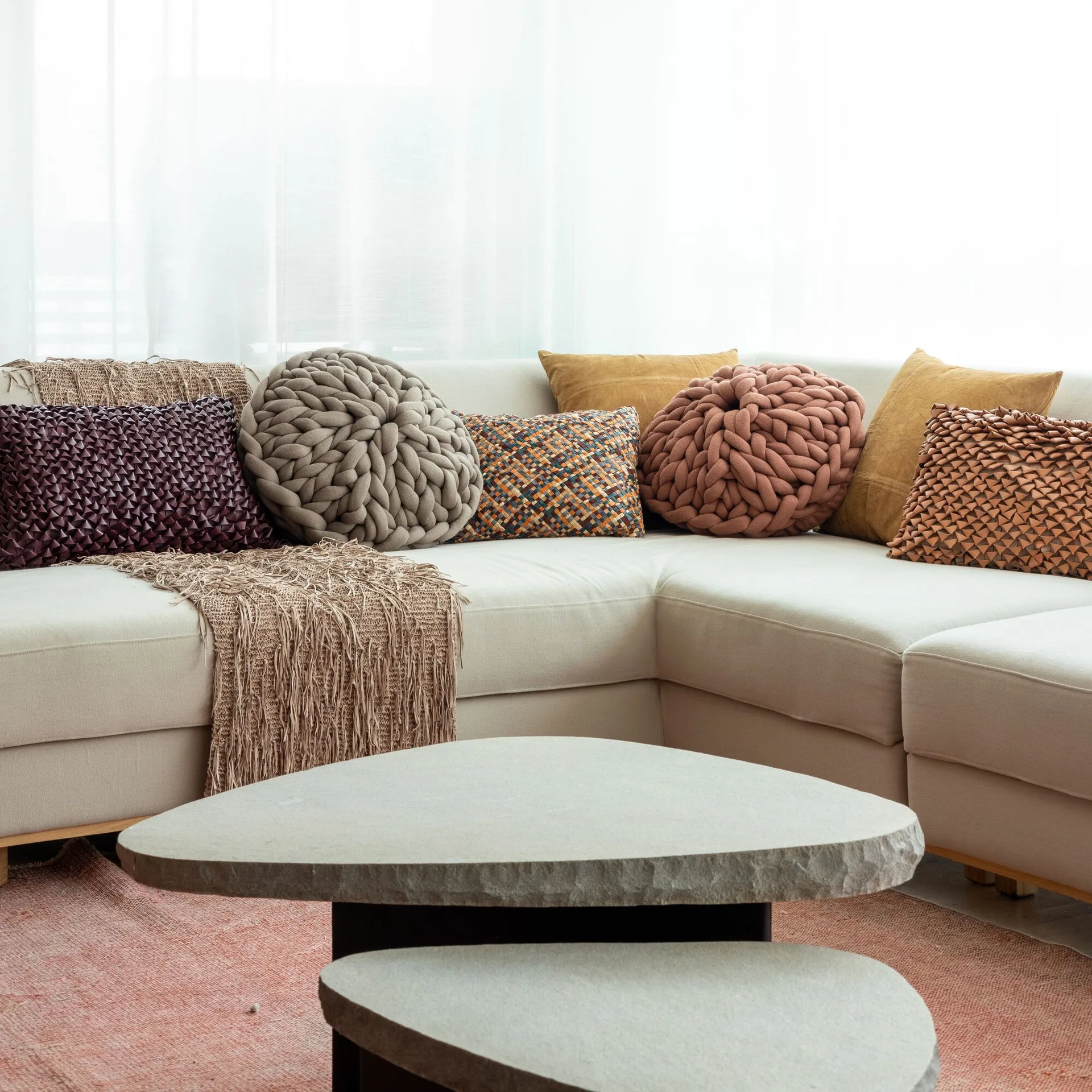 A cream sectional sofa layered with textured pillows sits behind two organic-shaped stone coffee tables on a terracotta rug.
