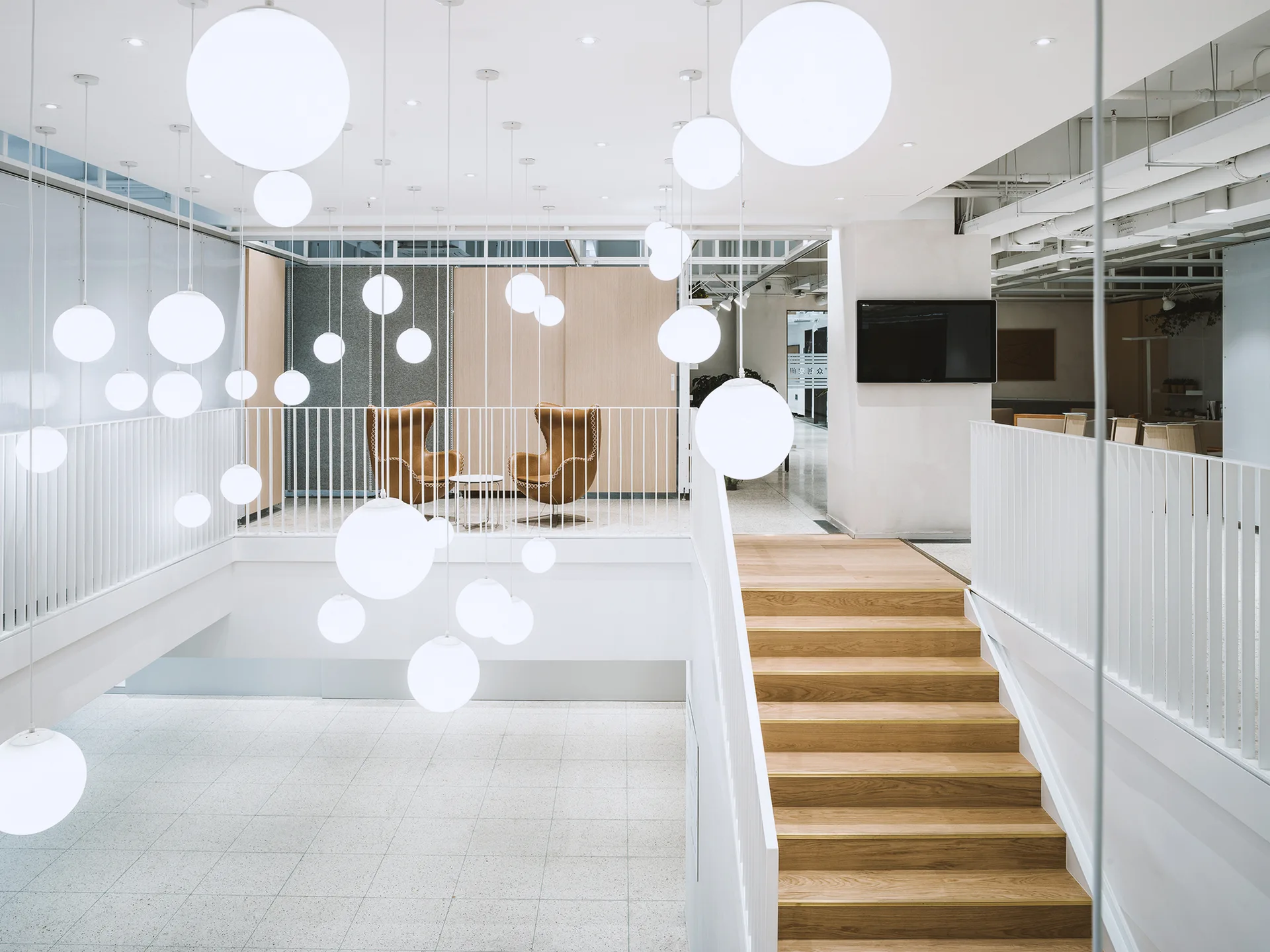 A modern white staircase with wooden steps leads to a mezzanine landing with two tan chairs under a cluster of suspended globe lights.