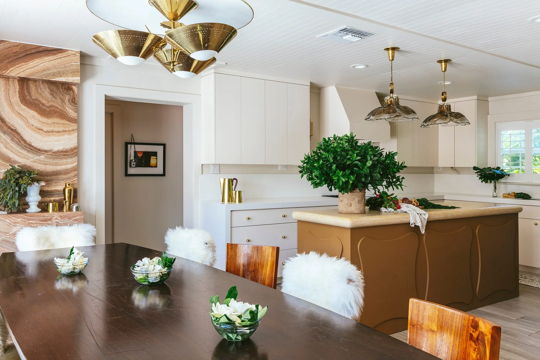An open-concept kitchen and dining area features a dark wood table, white shaggy chair covers, a brown kitchen island, and brass pendant lights.