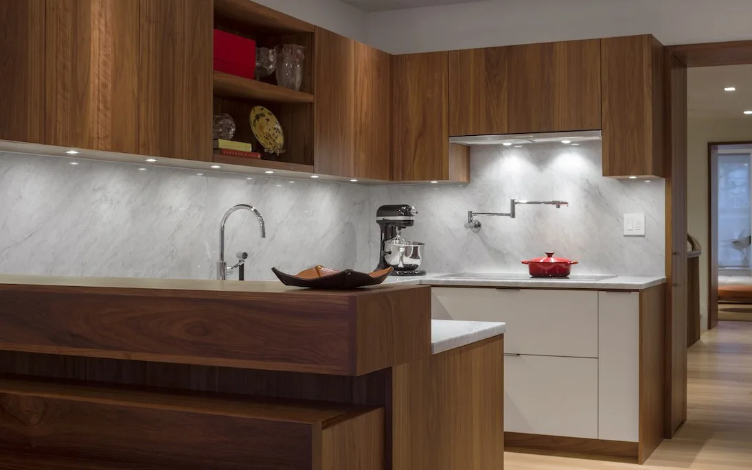 A modern kitchen features rich walnut upper cabinets, a veined grey-and-white marble slab backsplash, and clean white lower cabinetry.