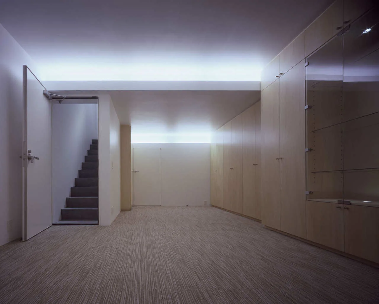 A minimalist hallway features a wall of light wood built-in cabinets, a patterned carpeted floor, indirect cove lighting, and a staircase.