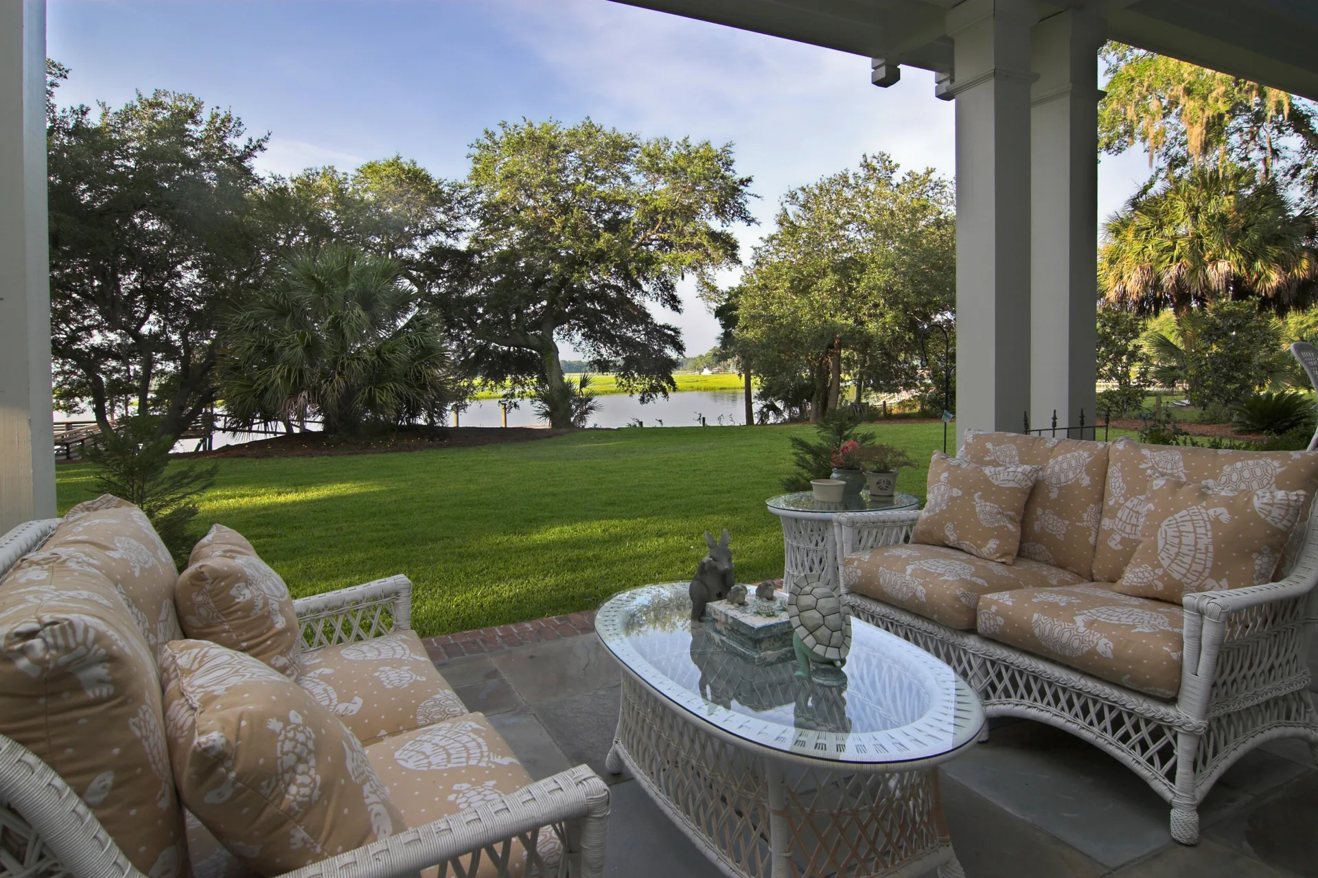 A covered porch featuring white wicker sofas with turtle-patterned tan cushions and glass-topped tables overlooking a grassy lawn and river.