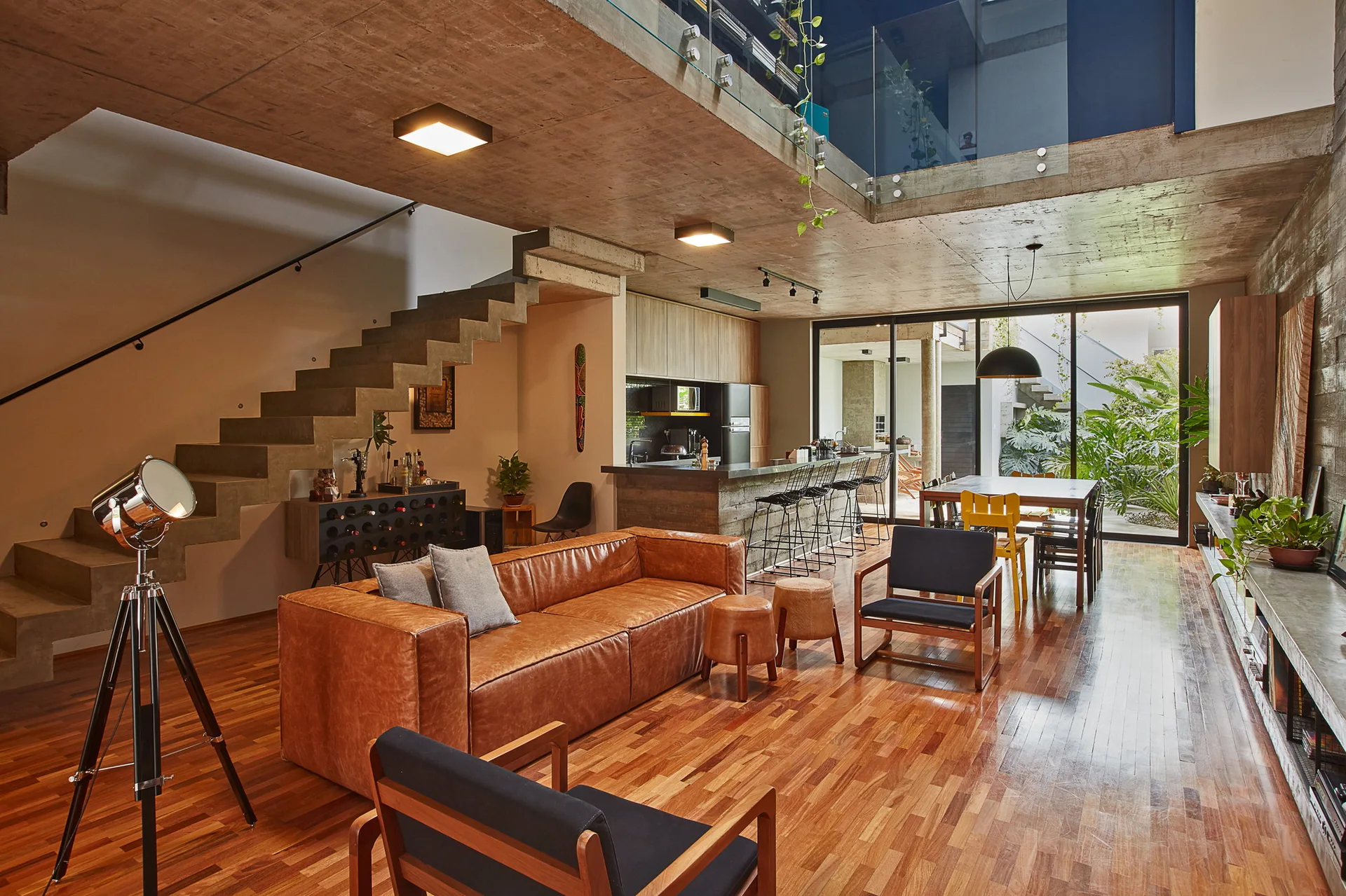 An open-plan living room features a tan leather sofa, concrete floating stairs, wood floors, and an exposed concrete ceiling.