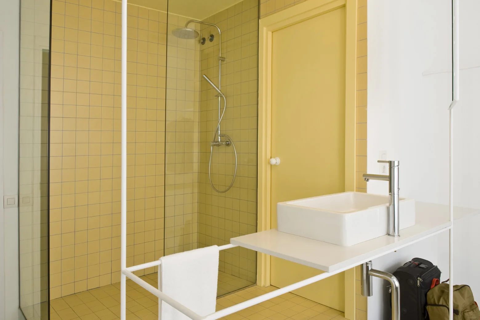 A minimalist bathroom features yellow square-tiled walls and floors, a clear glass shower, a yellow door, and a white vanity with a vessel sink.