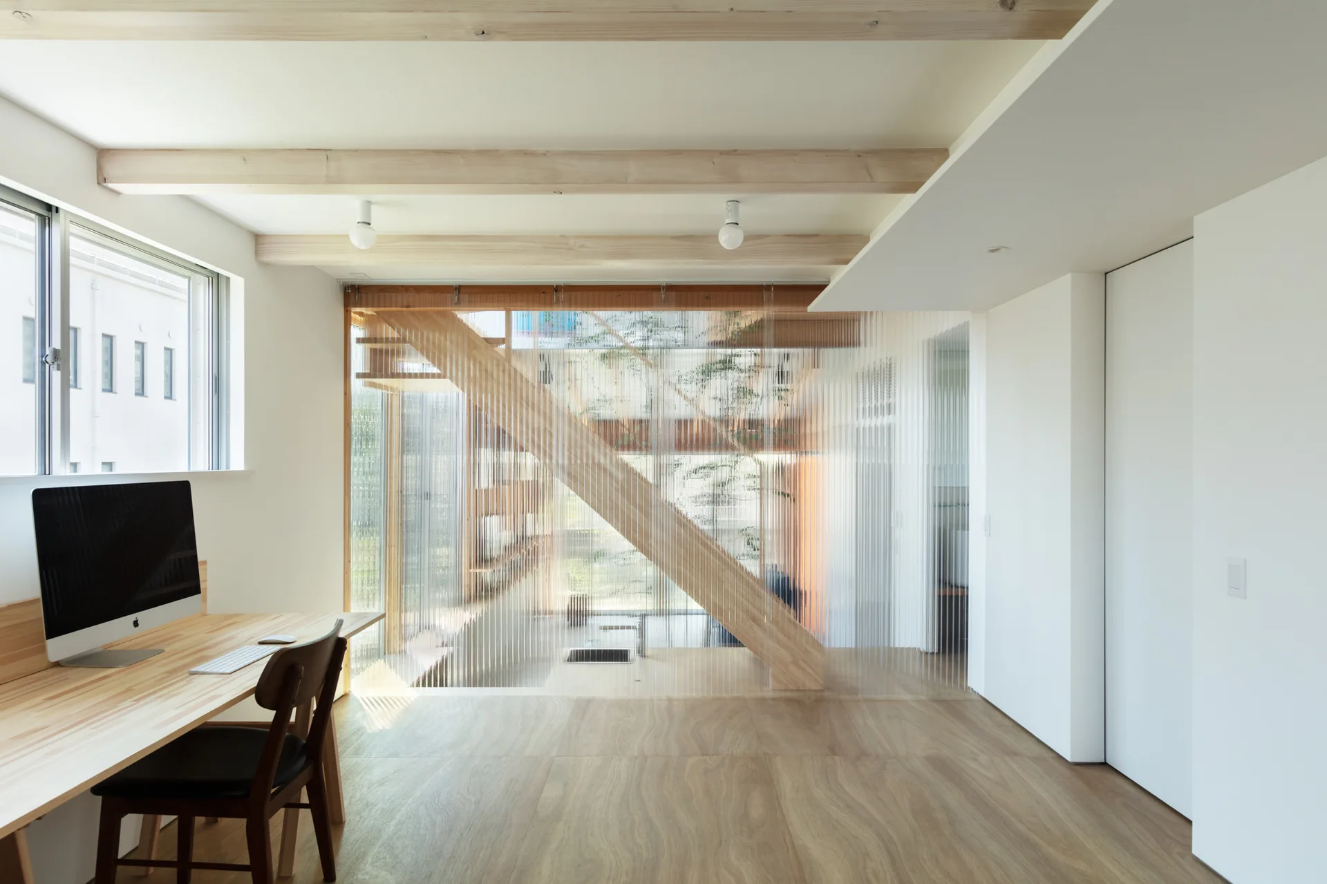A minimalist home office features a light wood desk, exposed ceiling beams, and a ribbed translucent partition wall overlooking a wooden staircase structure.