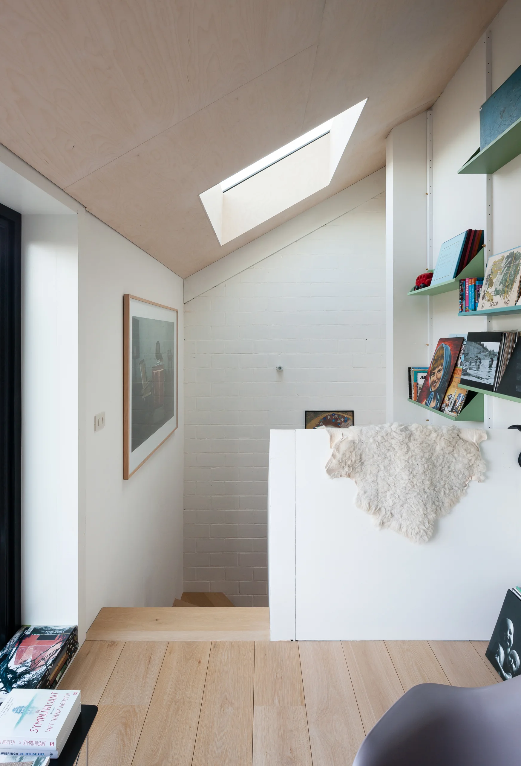A modern staircase landing features an angled plywood ceiling with a skylight, white painted brick walls, and pale green built-in shelves.