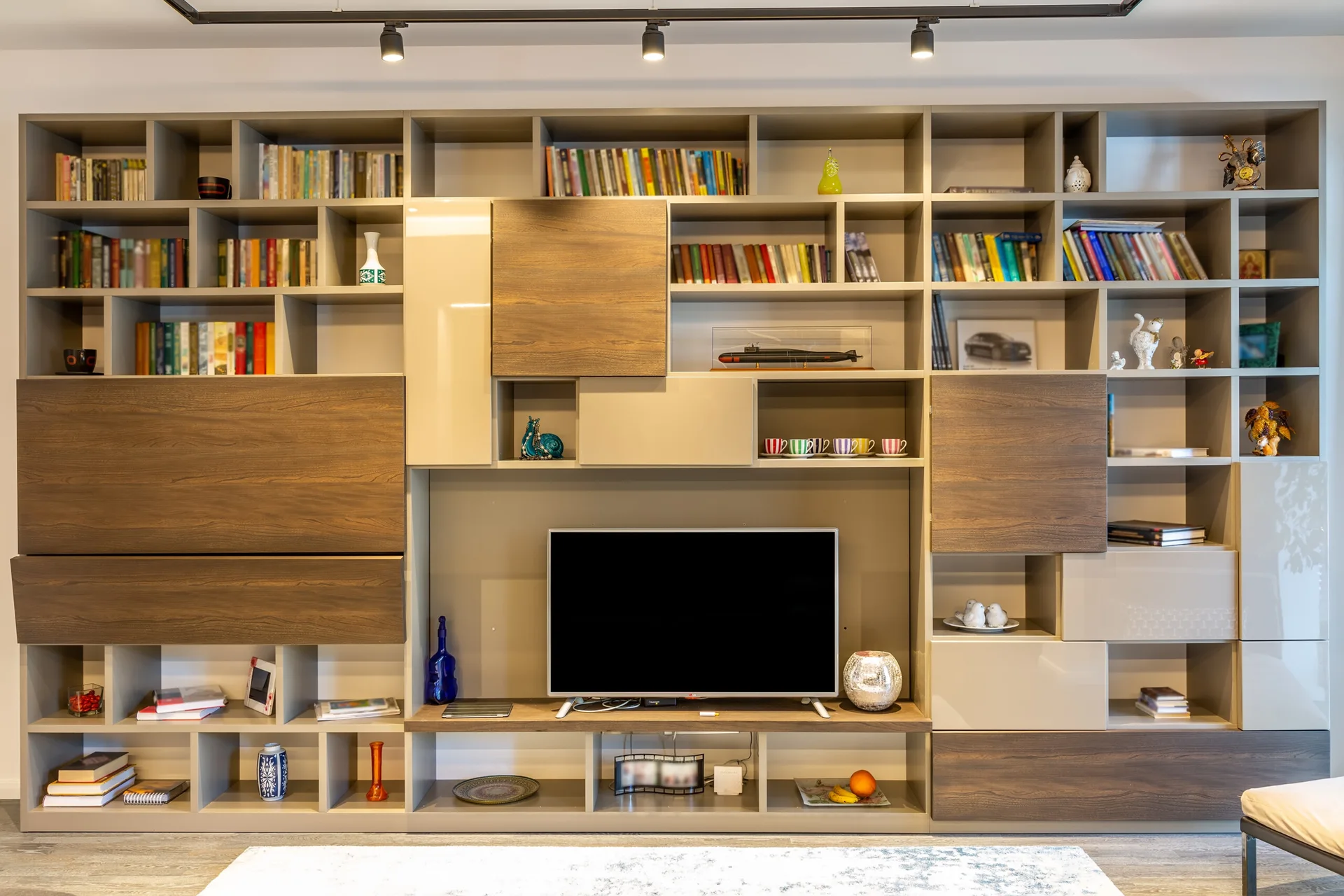 A large custom built-in media wall features asymmetrical shelves, wood grain cabinets, and a flat-screen television under track lighting.
