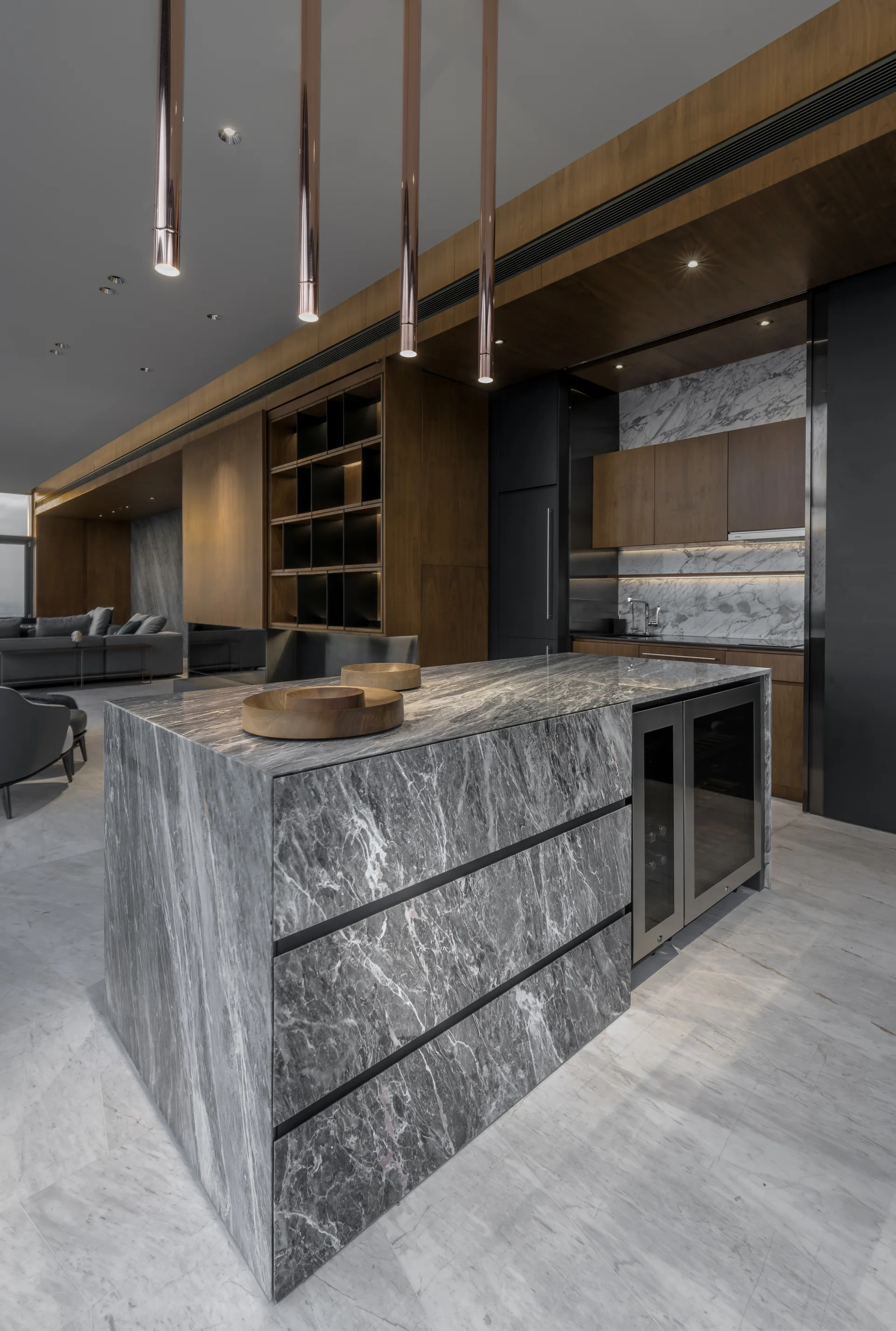 A modern kitchen showcasing a large grey veined marble island with a built-in wine cooler, wood cabinetry, and slim copper pendant lights.