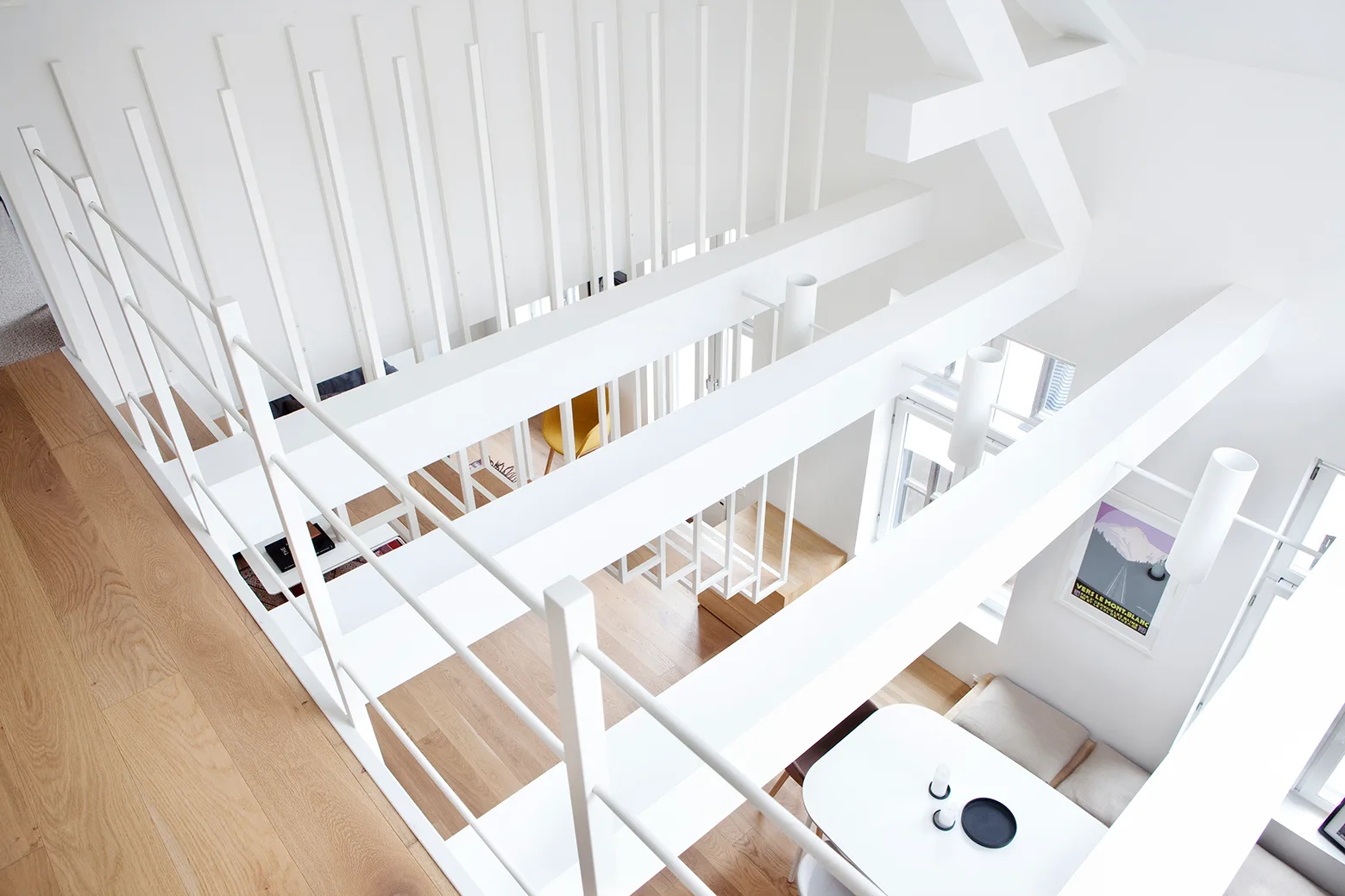 A high-angle view looking down from a white wood-floored landing with minimalist railings onto a dining area beneath large structural beams.