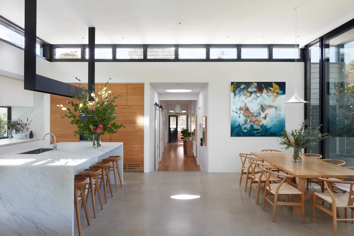 A modern open-plan kitchen and dining area with a marble waterfall island, wood cabinets, a wooden dining table, and high clerestory windows.