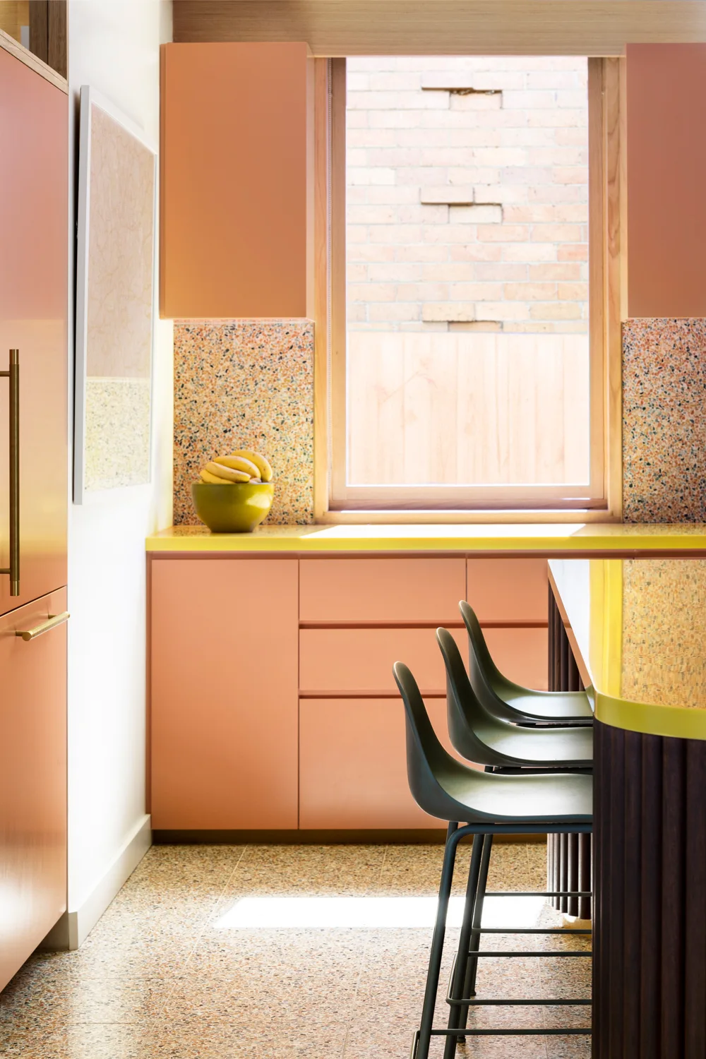 A contemporary kitchen featuring peach-colored cabinets, a curved island with dark green bar stools, and a speckled terrazzo backsplash and floor.