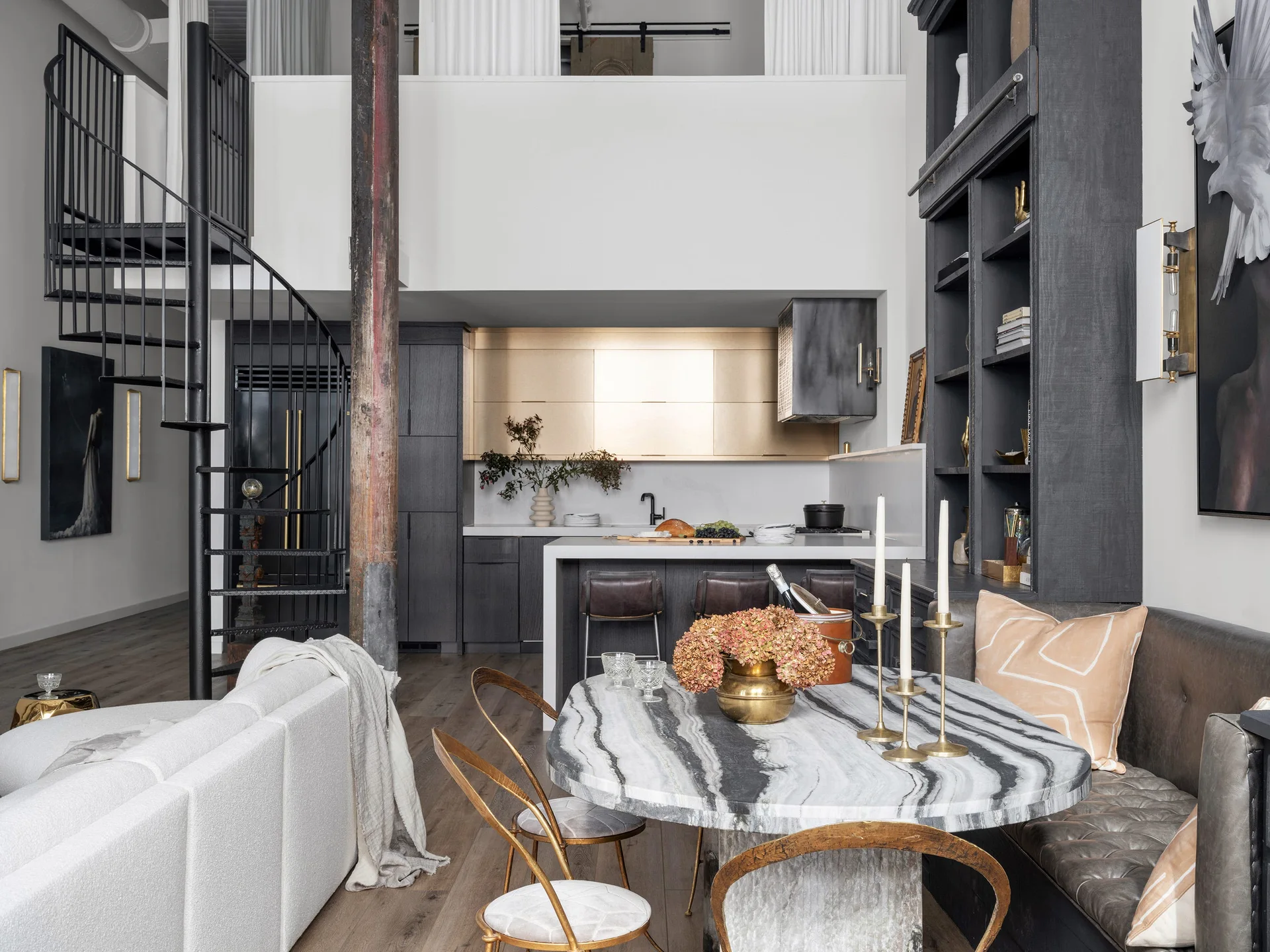 An open-plan loft features a heavily veined marble dining table with gold chairs, a black spiral staircase, and a kitchen under a mezzanine.