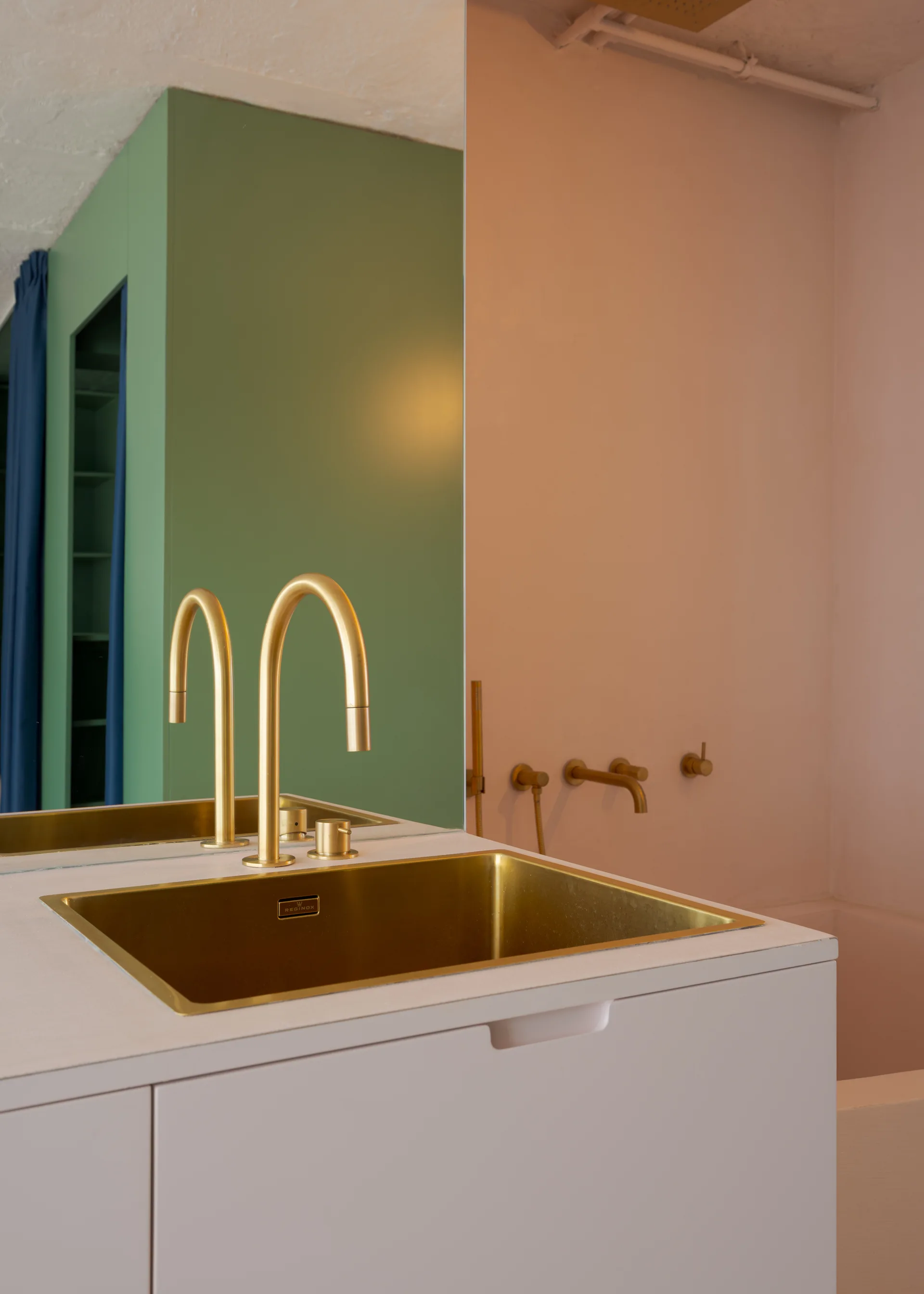 A brushed brass rectangular sink and matching arch faucet are set into a white vanity cabinet against pink and green color-blocked walls.