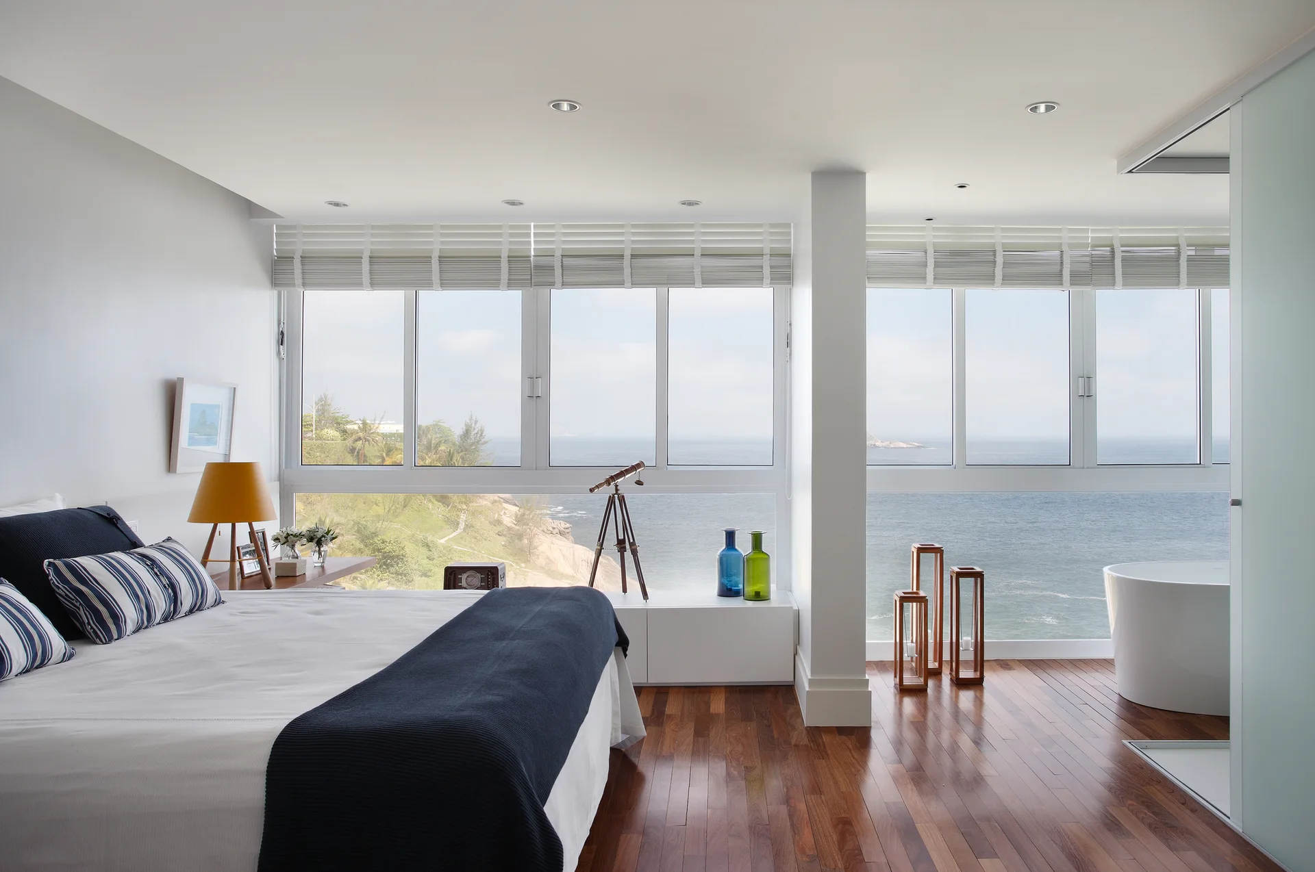 A bright coastal bedroom with a navy and white bed, a freestanding bathtub in the corner, and large windows overlooking the ocean.