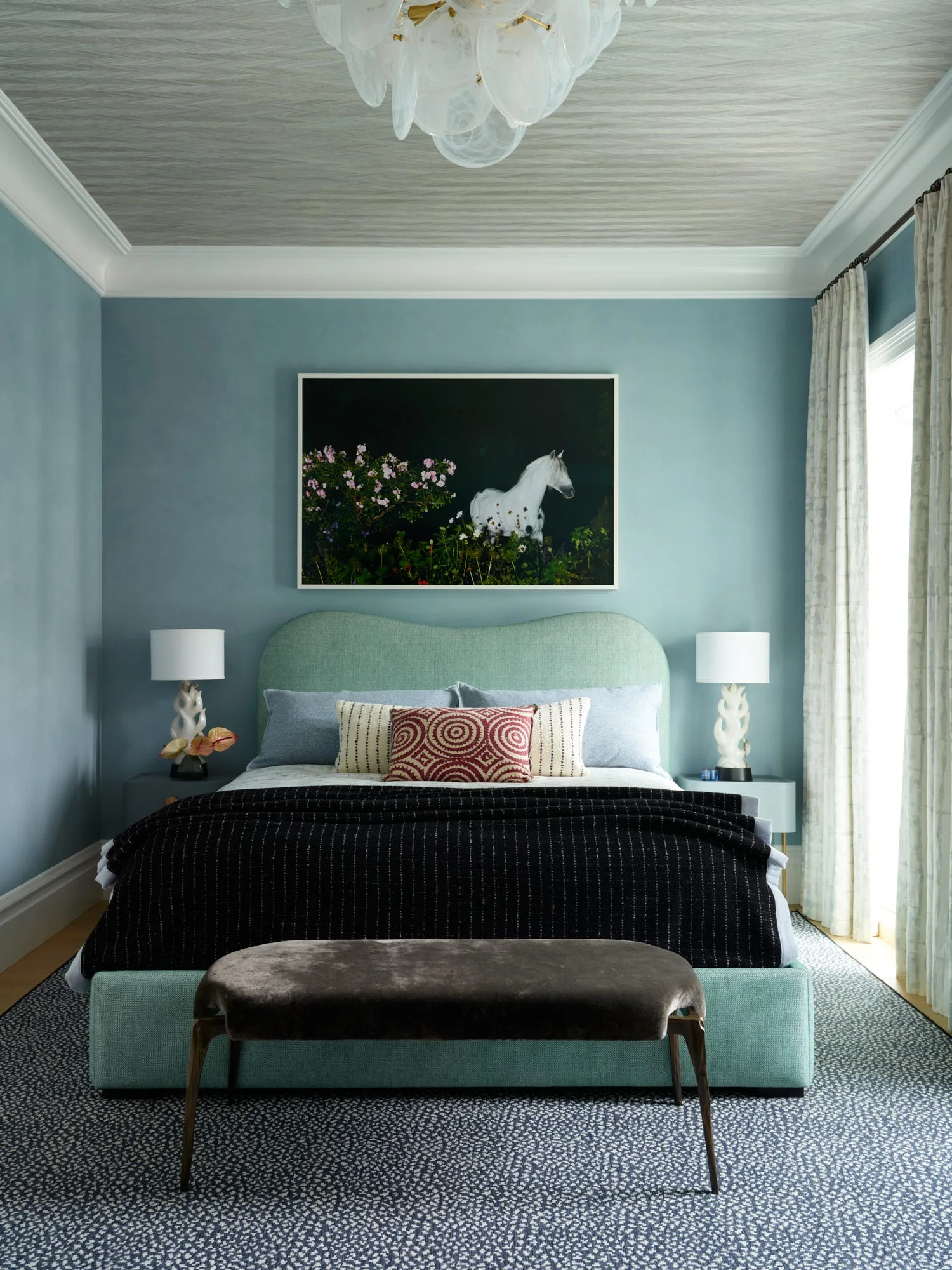 A bedroom with blue walls features a light green upholstered bed, a patterned ceiling, sculptural white bedside lamps, and a dark velvet bench.