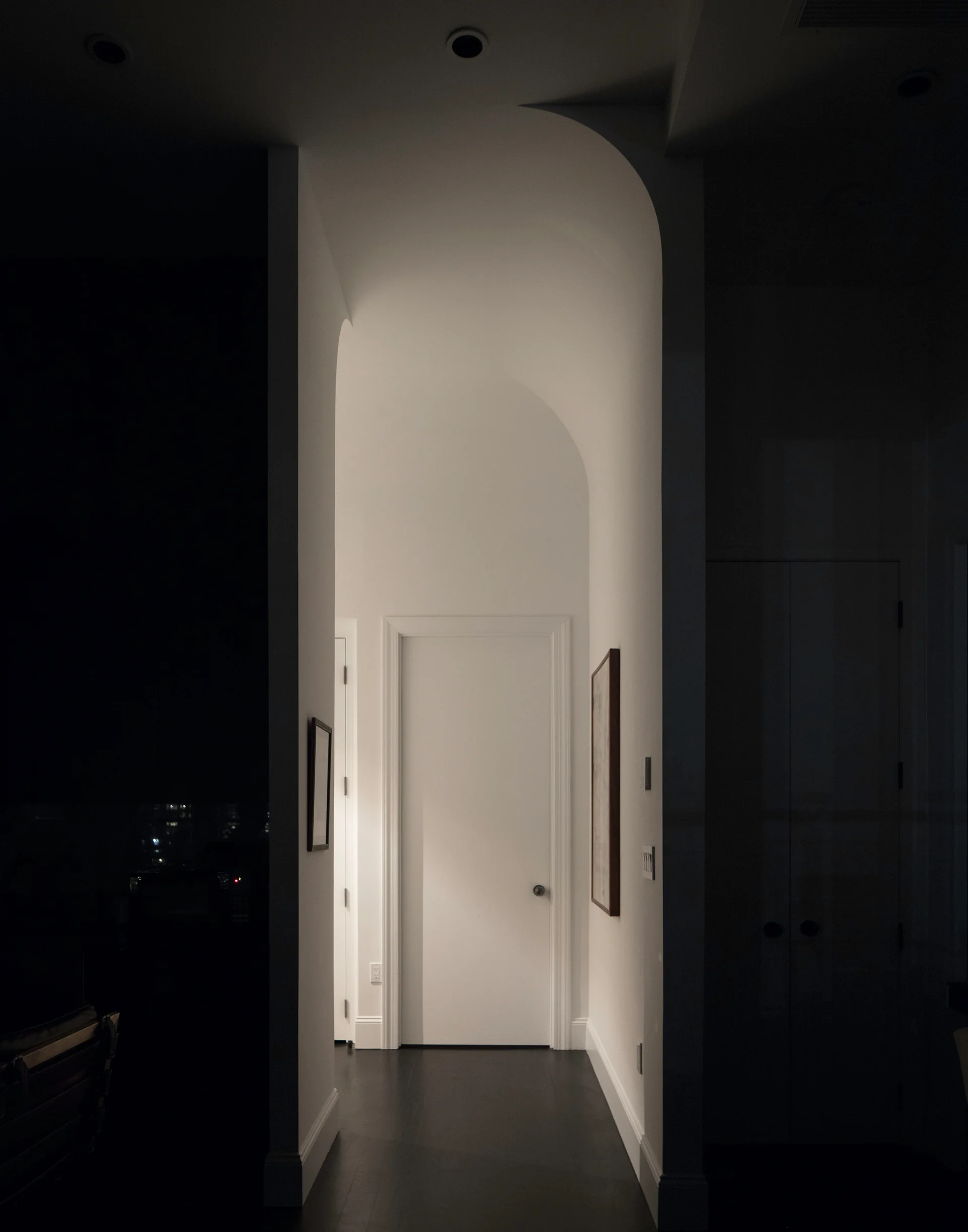 A narrow white hallway featuring a high arched ceiling, dark wood floors, and a white door framed by dark shadowed walls on either side.