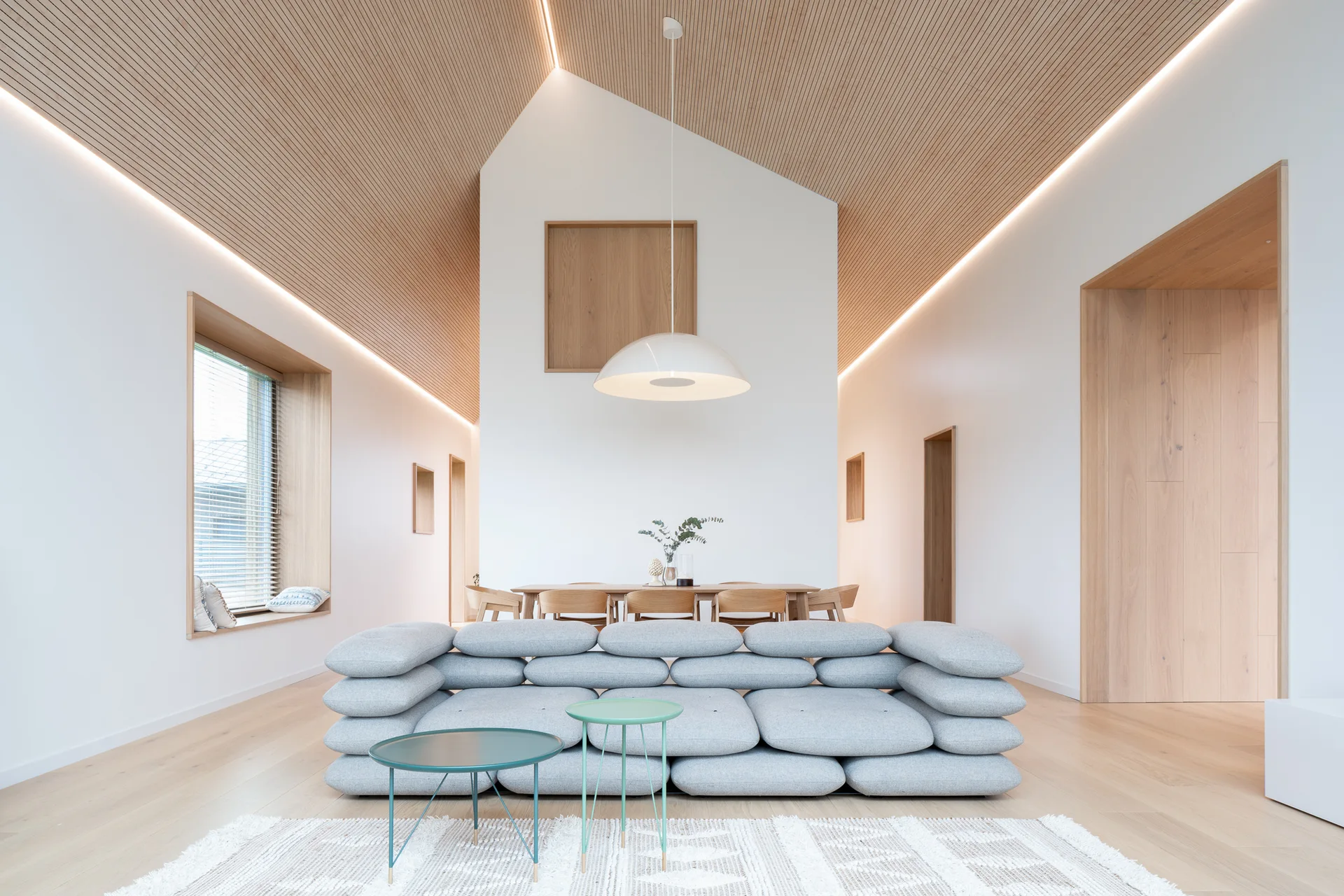 An open-plan living room featuring a vaulted wood-slat ceiling, a grey stacked-pillow sofa, and a dining table under a dome pendant.