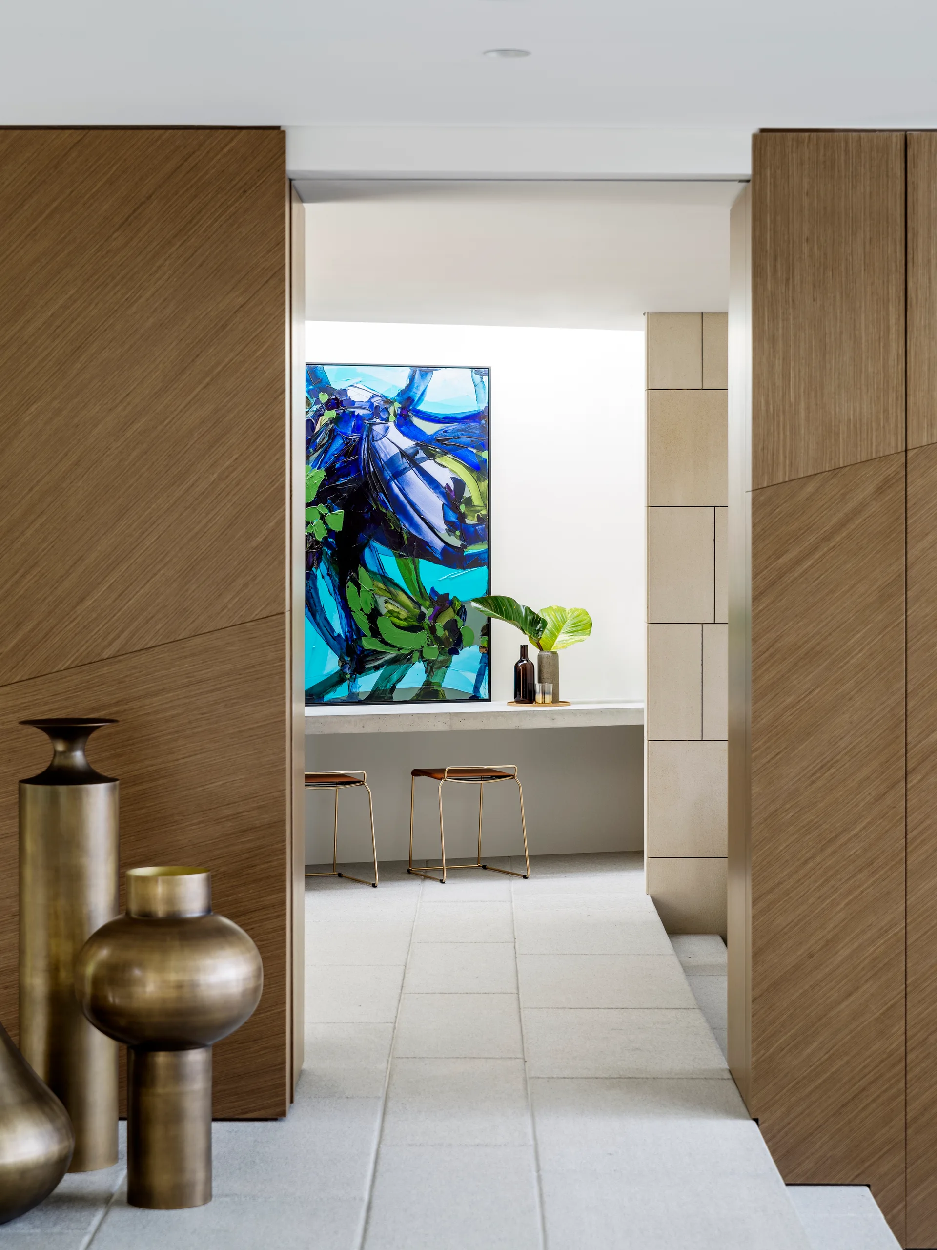 A transitional hallway framed by diagonal wood paneling features bronze floor vases in the foreground and a blue abstract painting in a bright background.