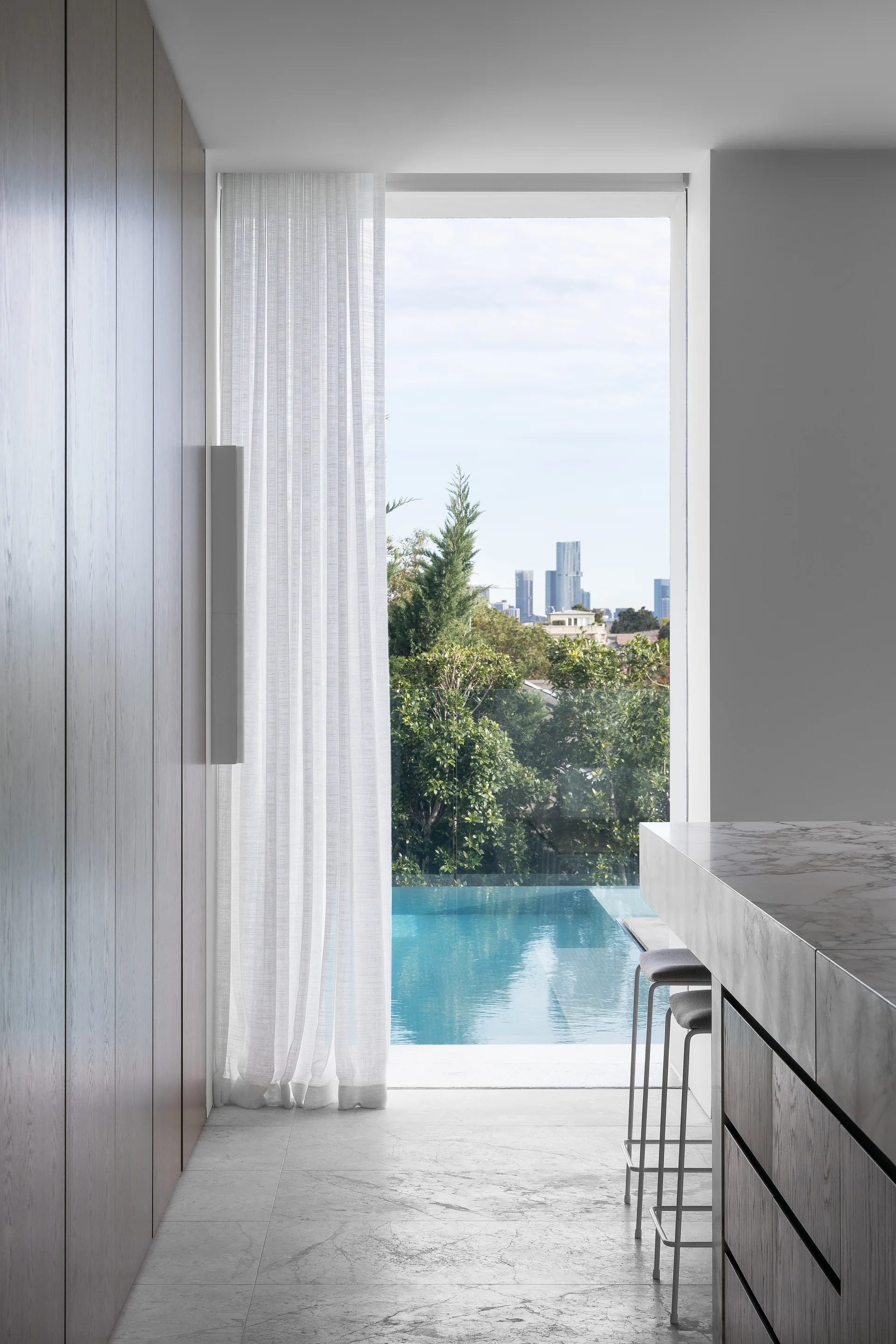 A minimalist kitchen features a marble-topped island with stools next to a tall glass window overlooking a blue swimming pool.