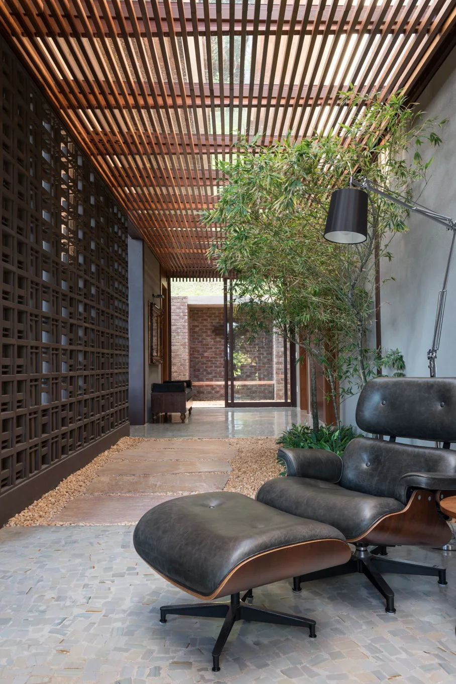 A covered outdoor patio features a black leather lounge chair, a slatted wood ceiling, gravel flooring with stone pavers, and a breeze block wall.