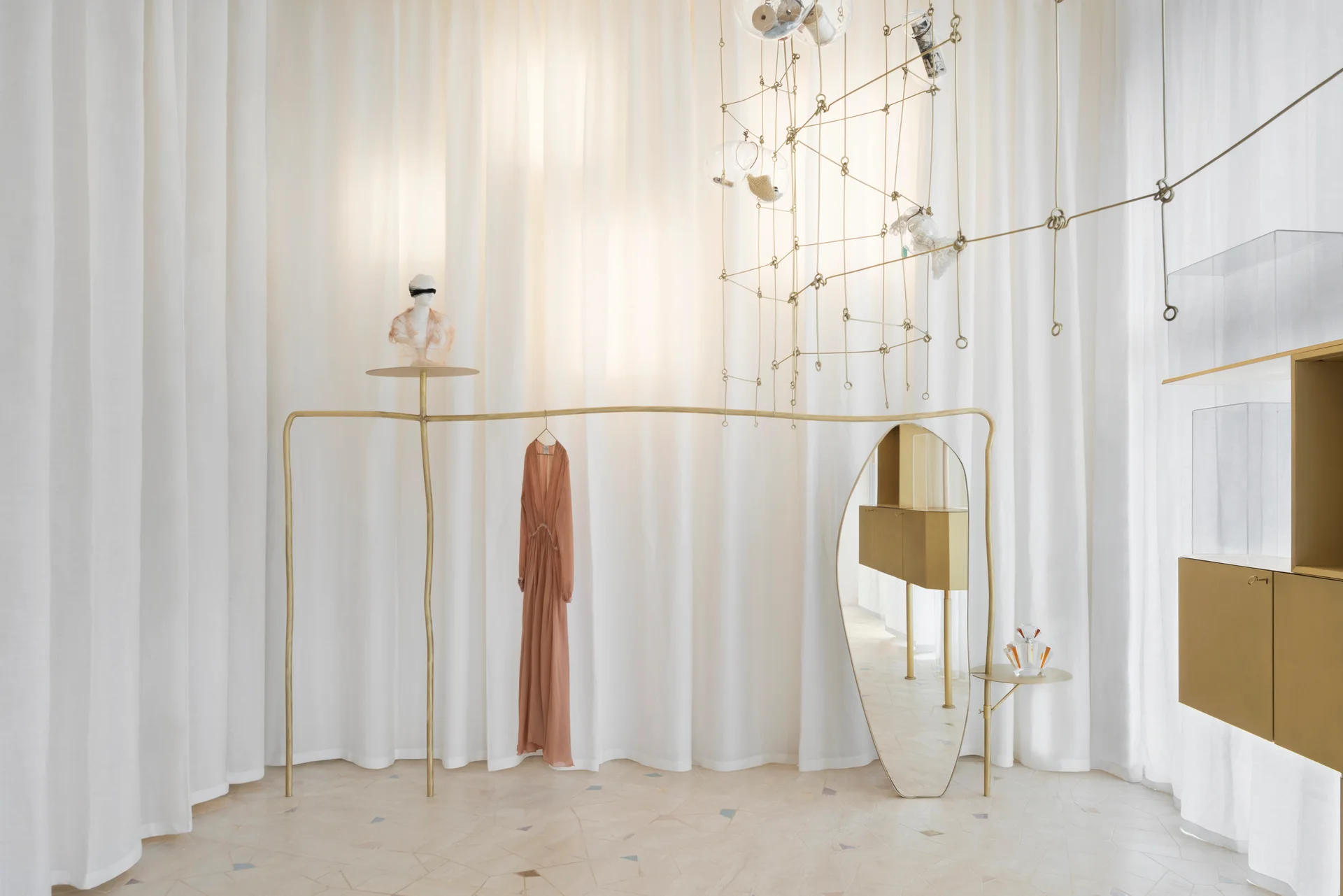 A sculptural brass clothing rack holding a peach dress stands against floor-to-ceiling white curtains next to an integrated oval mirror.