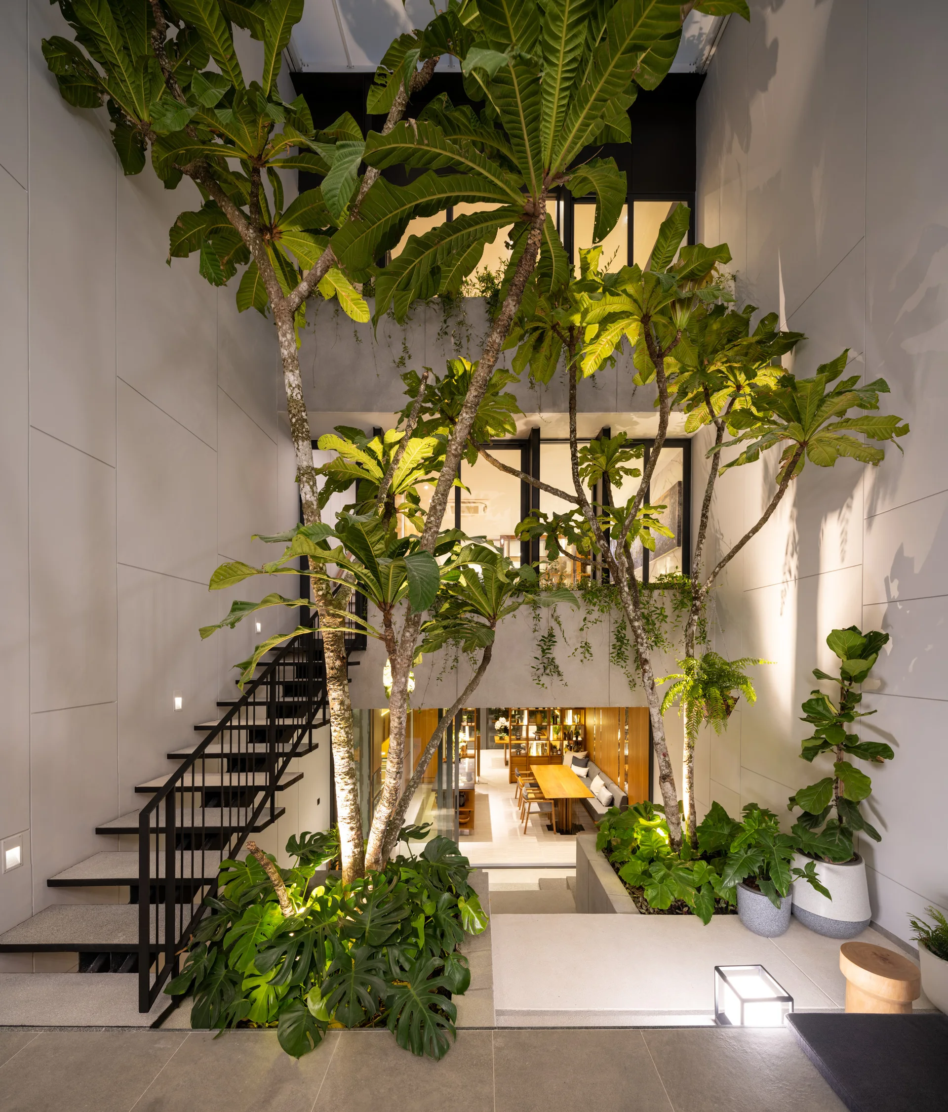 A modern multi-level courtyard features tall mature trees, integrated ground plantings, an open-riser black metal staircase, and concrete paneled walls.