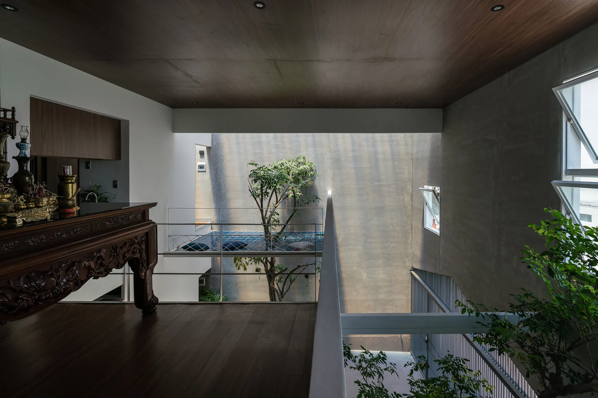 A modern indoor landing with wood flooring overlooks a concrete courtyard void containing a living tree and a suspended net hammock.