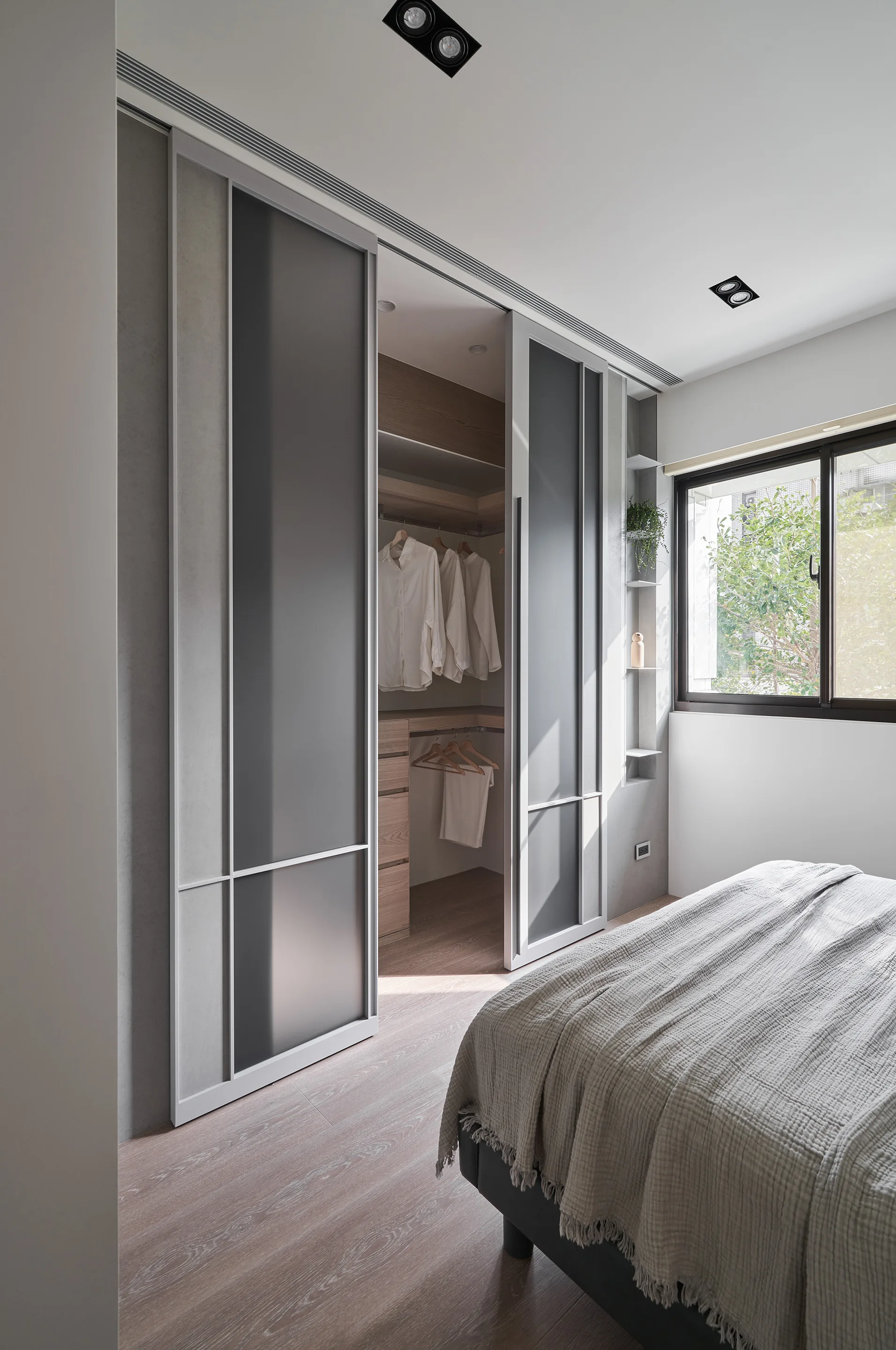 A modern bedroom features a walk-in wardrobe with frosted glass sliding doors next to a bed with textured grey linens.