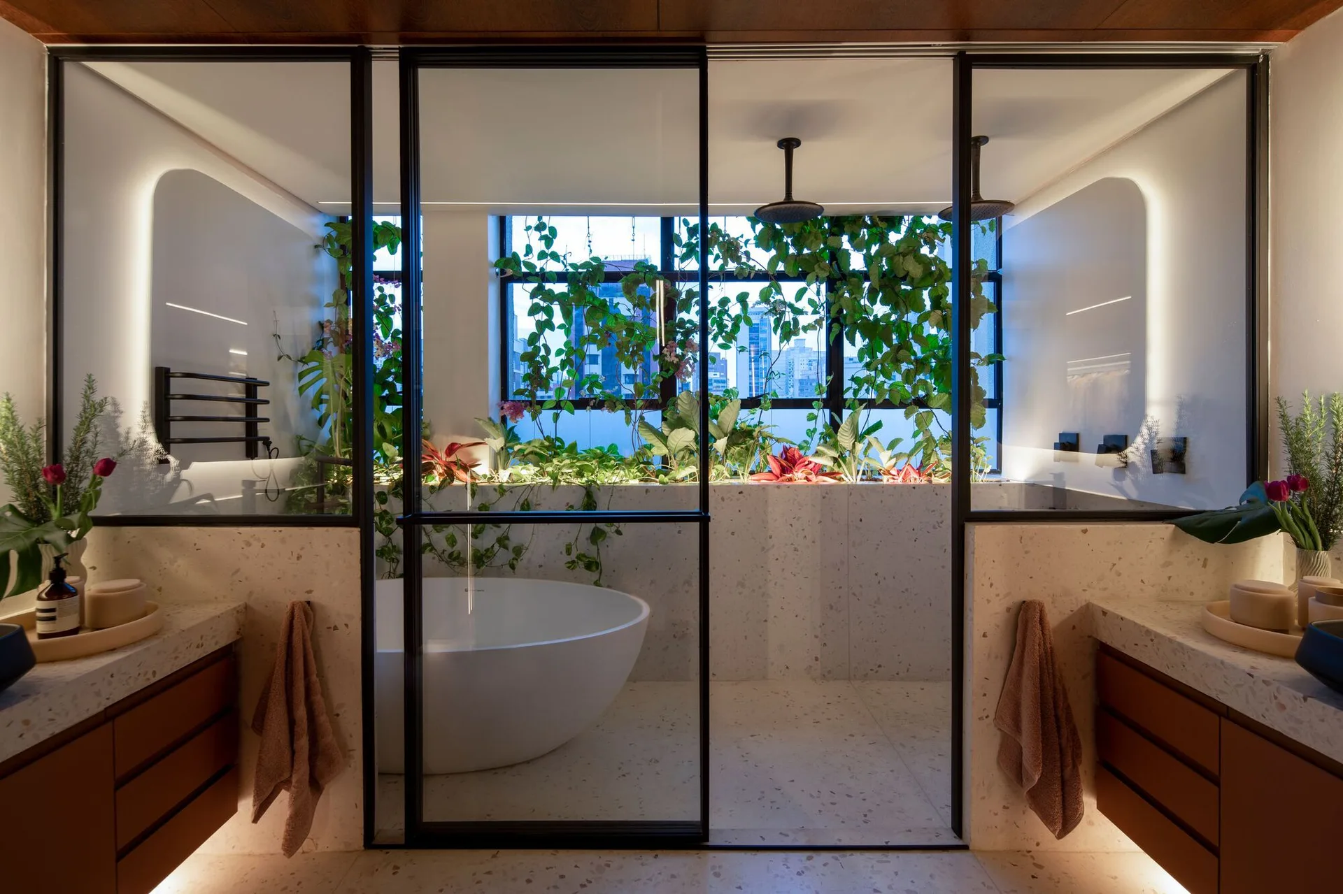 A modern bathroom with a freestanding white tub behind a black-framed glass divider and a lush plant-filled ledge against the window.
