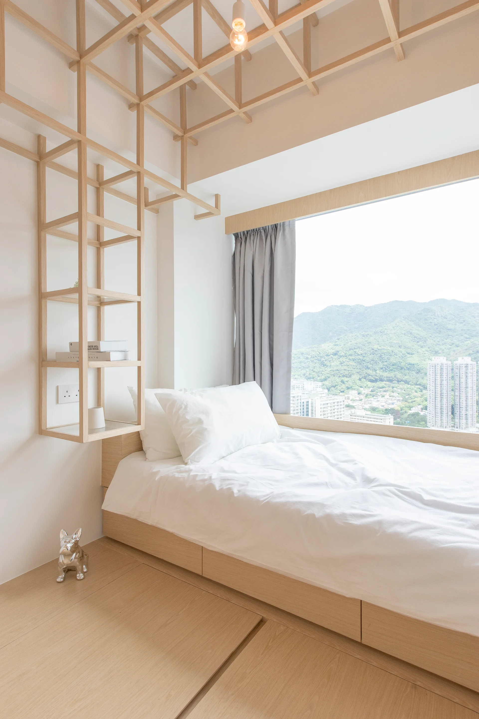 A light wood platform bed with white sheets sits beneath a decorative timber ceiling grid next to a large window overlooking green mountains.