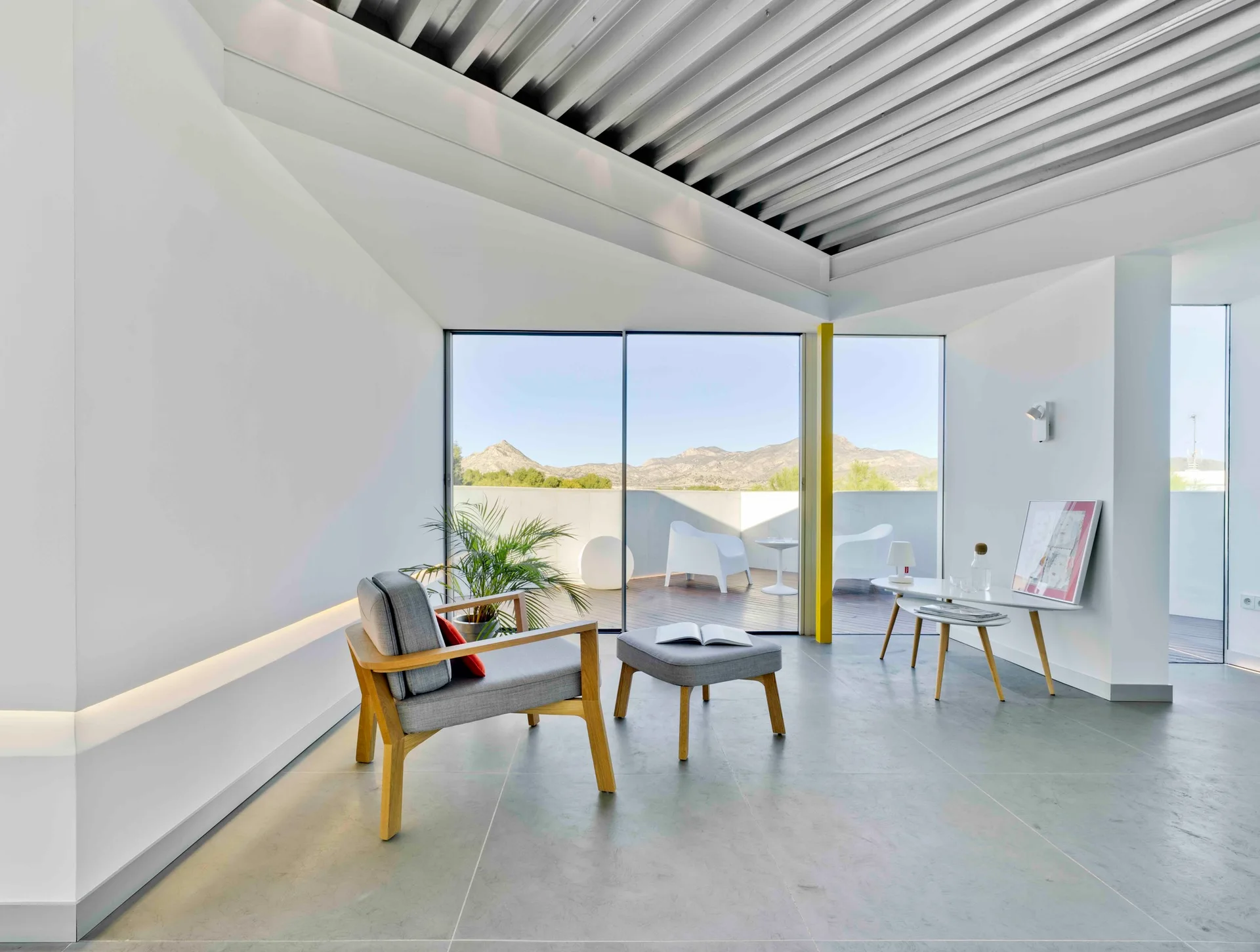 A minimalist living room with a grey fabric armchair, a corrugated metal ceiling, and large glass doors leading to an outdoor terrace.