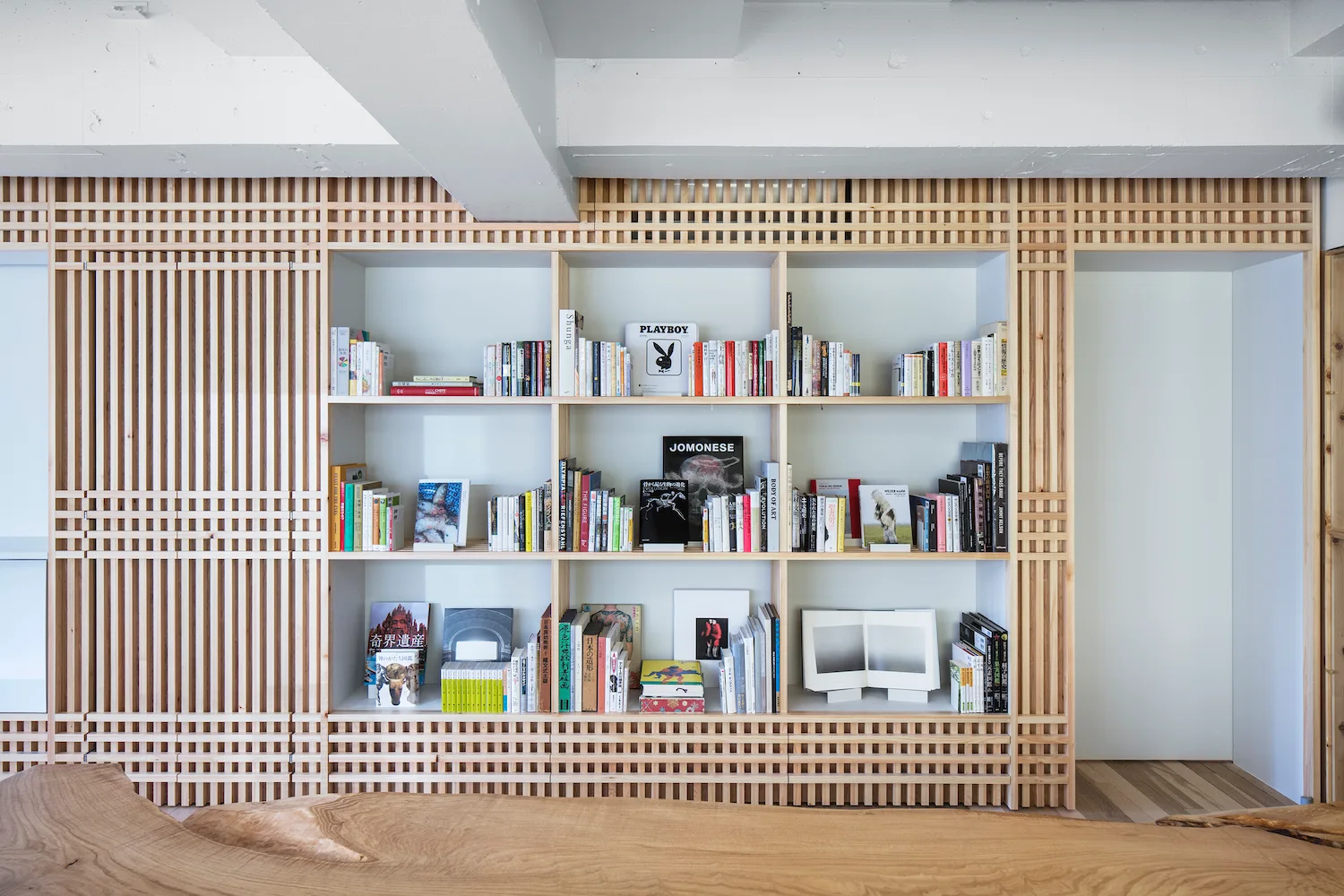 A large built-in wooden bookshelf filled with books, featuring light wood lattice paneling and set under an exposed concrete ceiling.