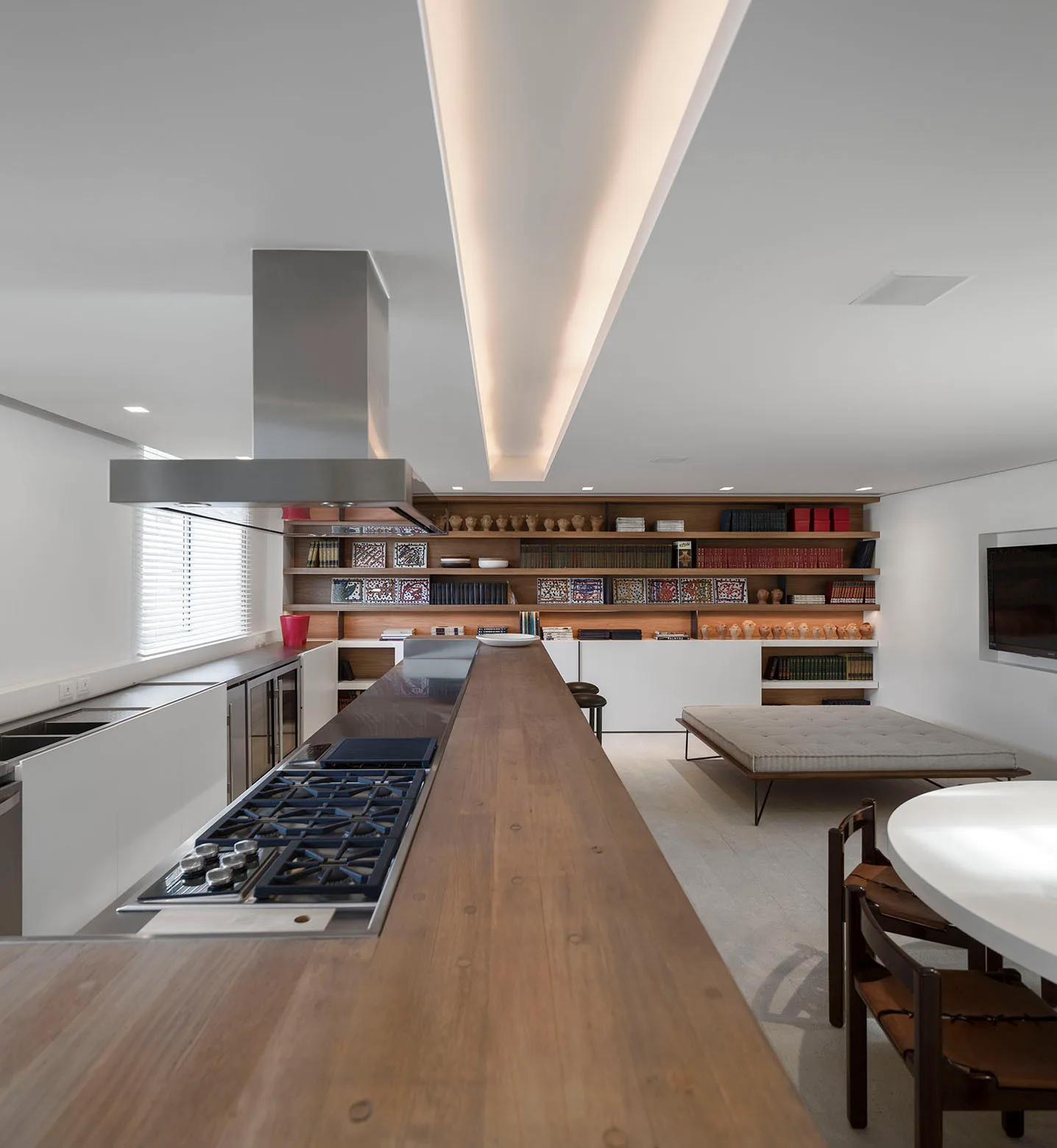 An open-plan kitchen featuring a long wooden island with a range hood, set against a large built-in wooden bookcase on the back wall.