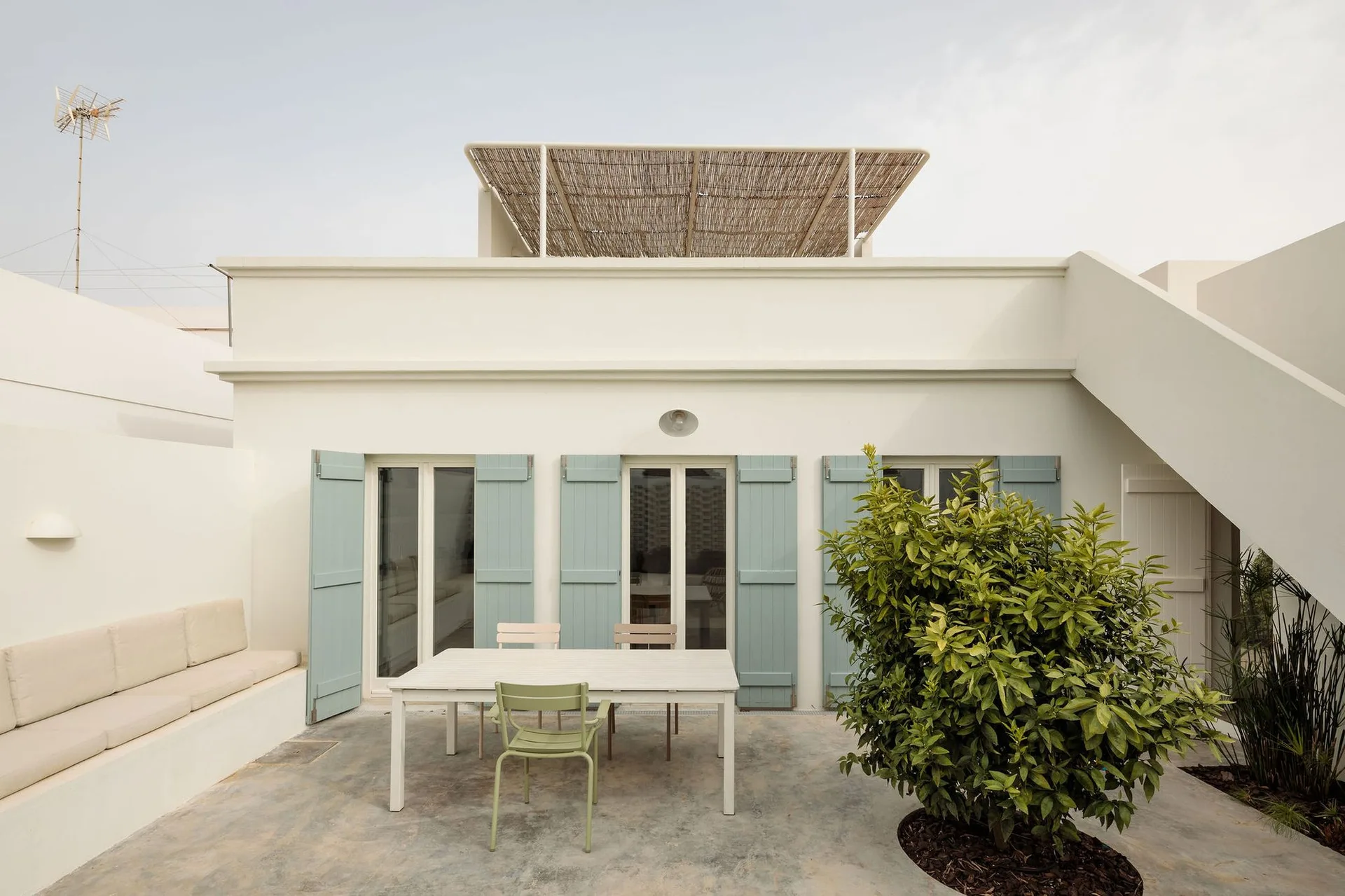 An outdoor white stucco patio features a dining table with chairs, light blue wooden shutters, and an integrated masonry bench.