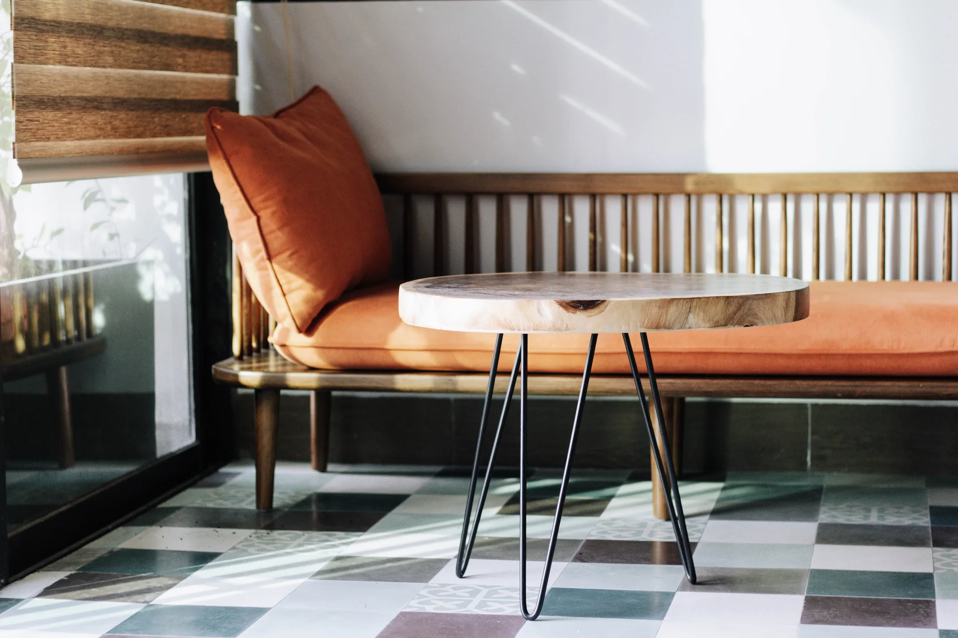 A wooden slatted bench with a burnt orange cushion stands on checkered floor tiles next to a round wood slab side table with black hairpin legs.
