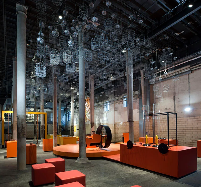 An industrial gallery space features concrete columns, a dark ceiling hung with wire cages, and bright orange display platforms.